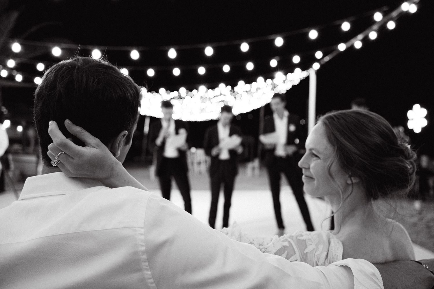 a black and white photo of a bride gently touching the back of the groom's head and looking at him lovingly as his groomsmen give a speech
