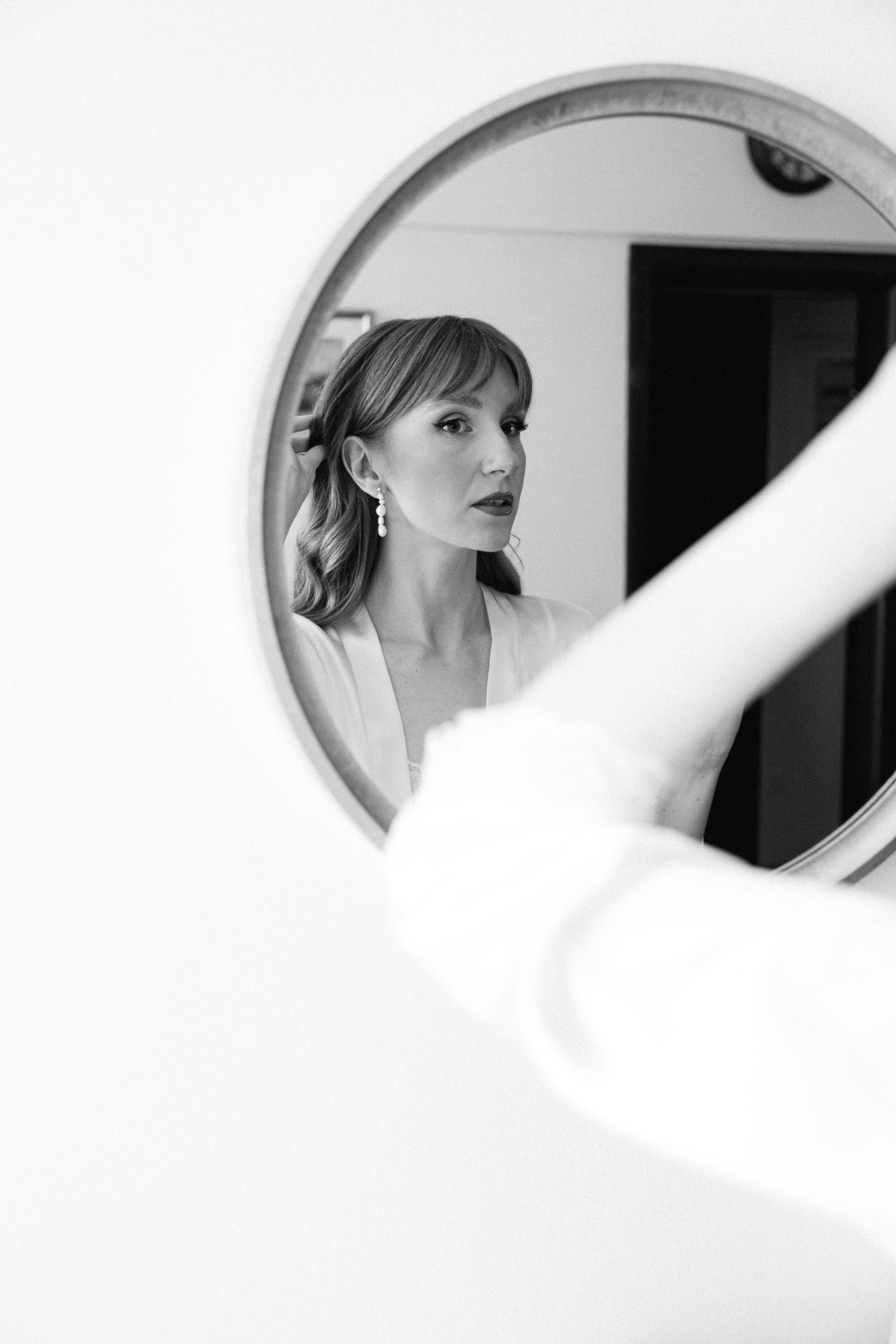 a black and white close up portrait of a bride looking at herself in the mirror fixing her hair, wearing dark lipstick