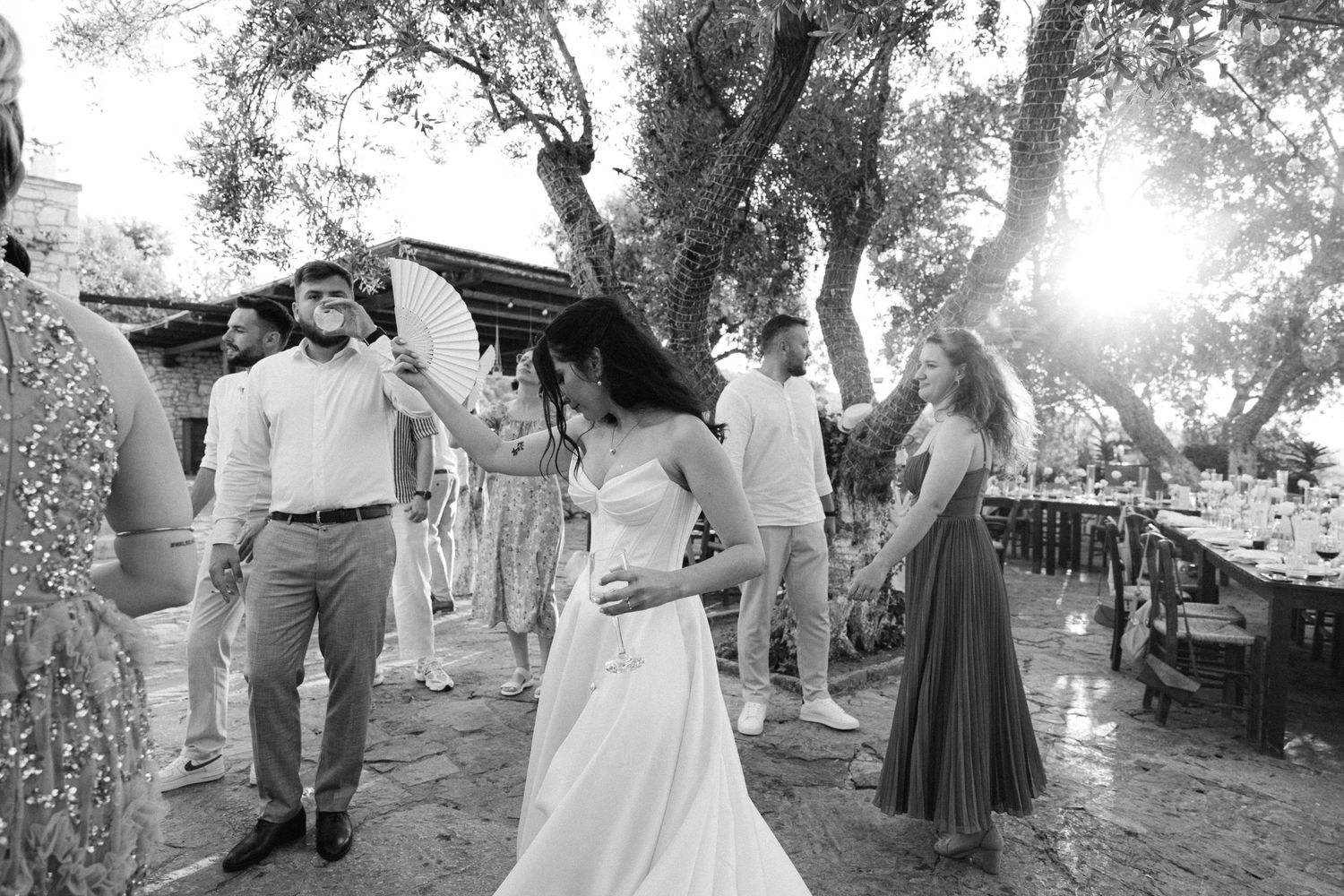a black and white photo of a party during a wedding reception - the guests and the bride are dancing at sunset in a nature rustic setting