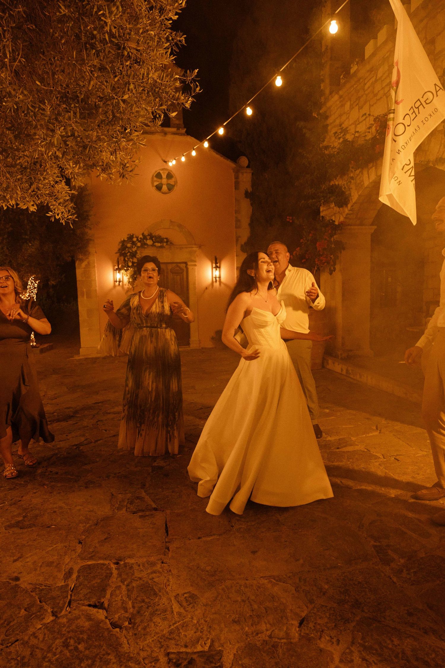 A bride dancing and singing loudly at night in front of a rustic chapel with tungsten lights with 3 more people dancing in the background