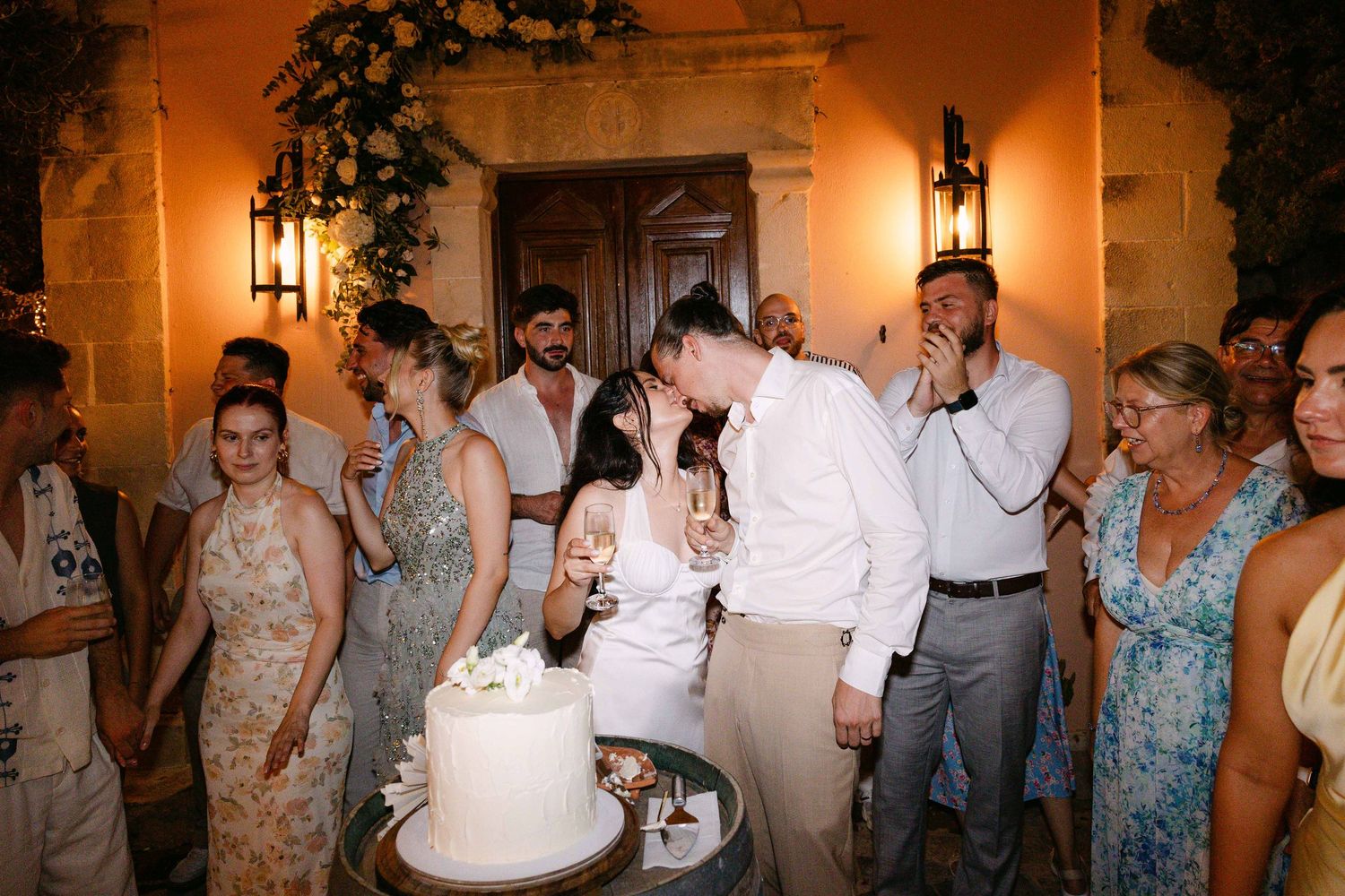 a bride and groom kissing while cutting their wedding cake surrounded by cheering guests at night in front of a chapel