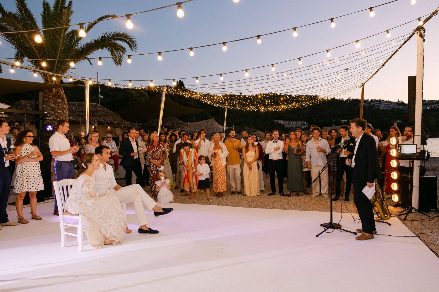 a couple sitting in chairs opposite the bride's dad who is reading a speech into a microphone with guests under carnival lights