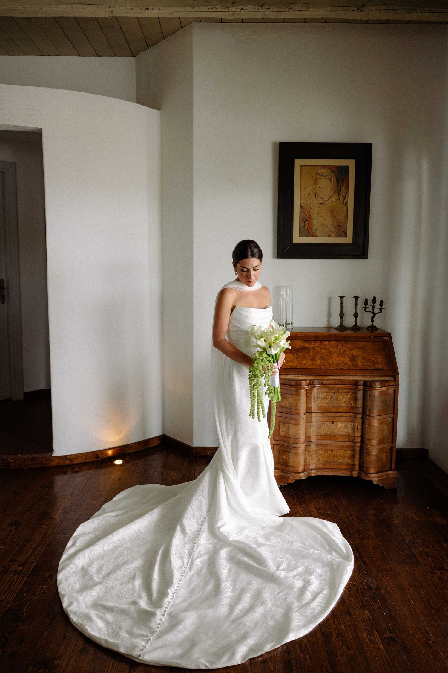a dim-lit portrait of a bride holding her bouquet of white Lillies and looking down standing in a antique-decorated living room