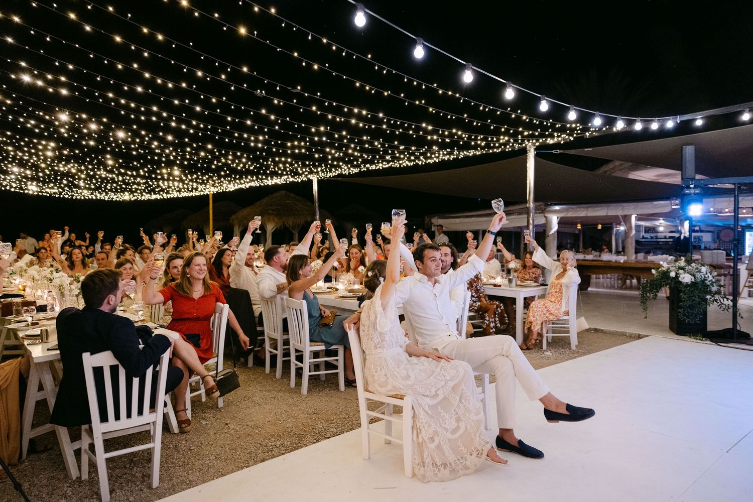 a bride and groom sitting in chairs and toasting with their wedding guests in an open-air wedding reception in Spetses