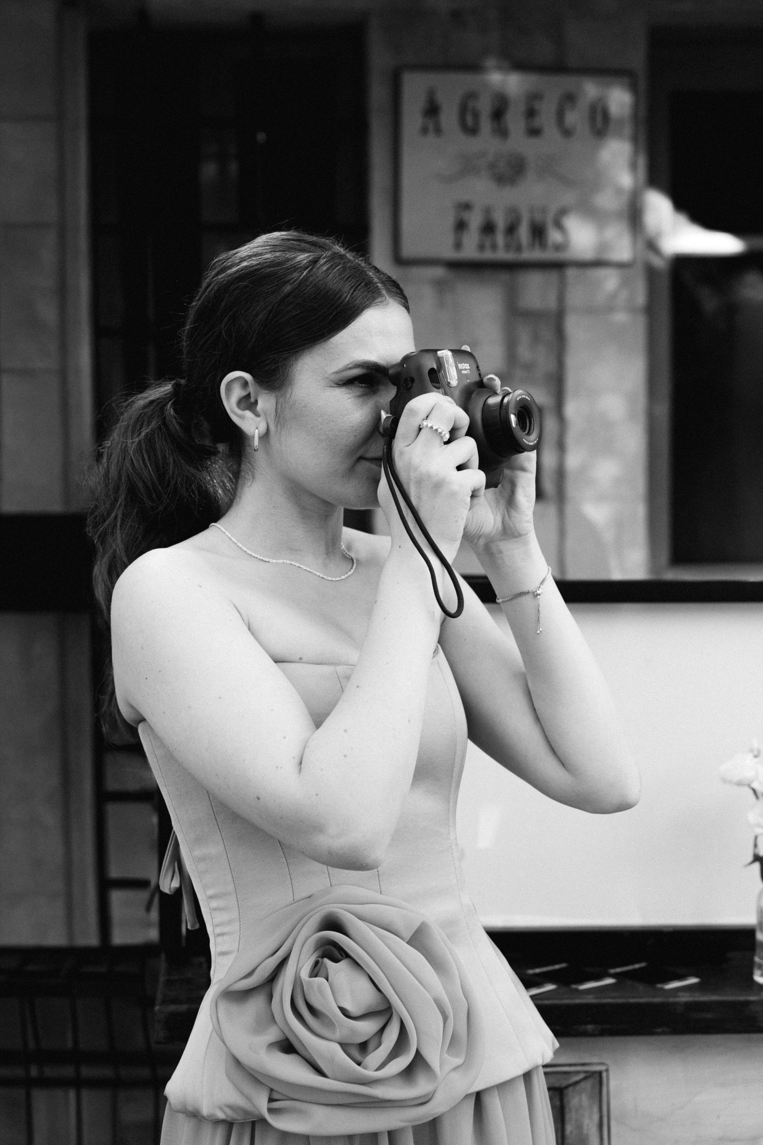 a black and white photo of a woman in a dress and a ponytail holding a camera and taking photos of something outside of our shot