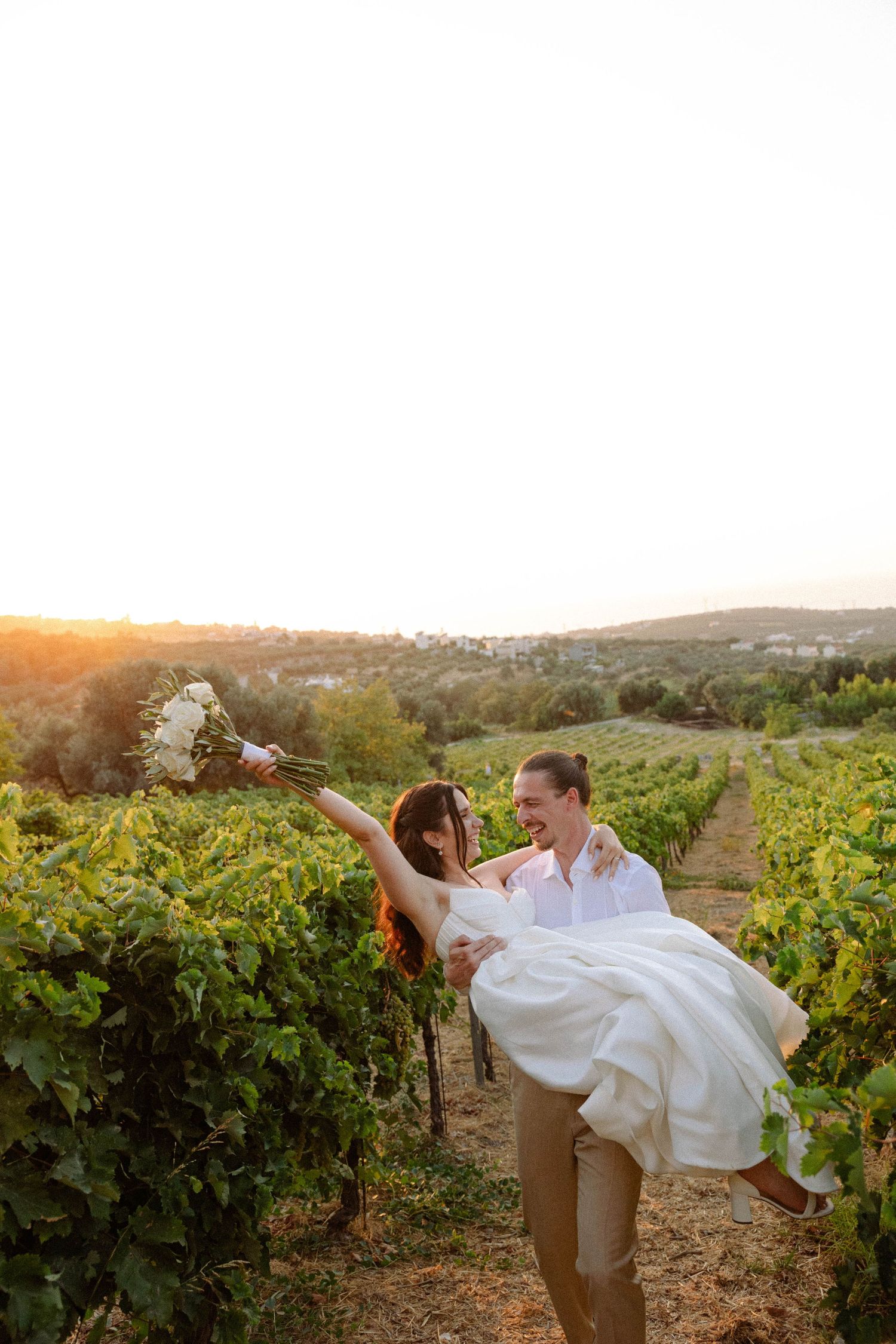 a groom holding and lifting a bride while she laughs and has her hands up in the air holding her white flowers bouquet in a vineyard