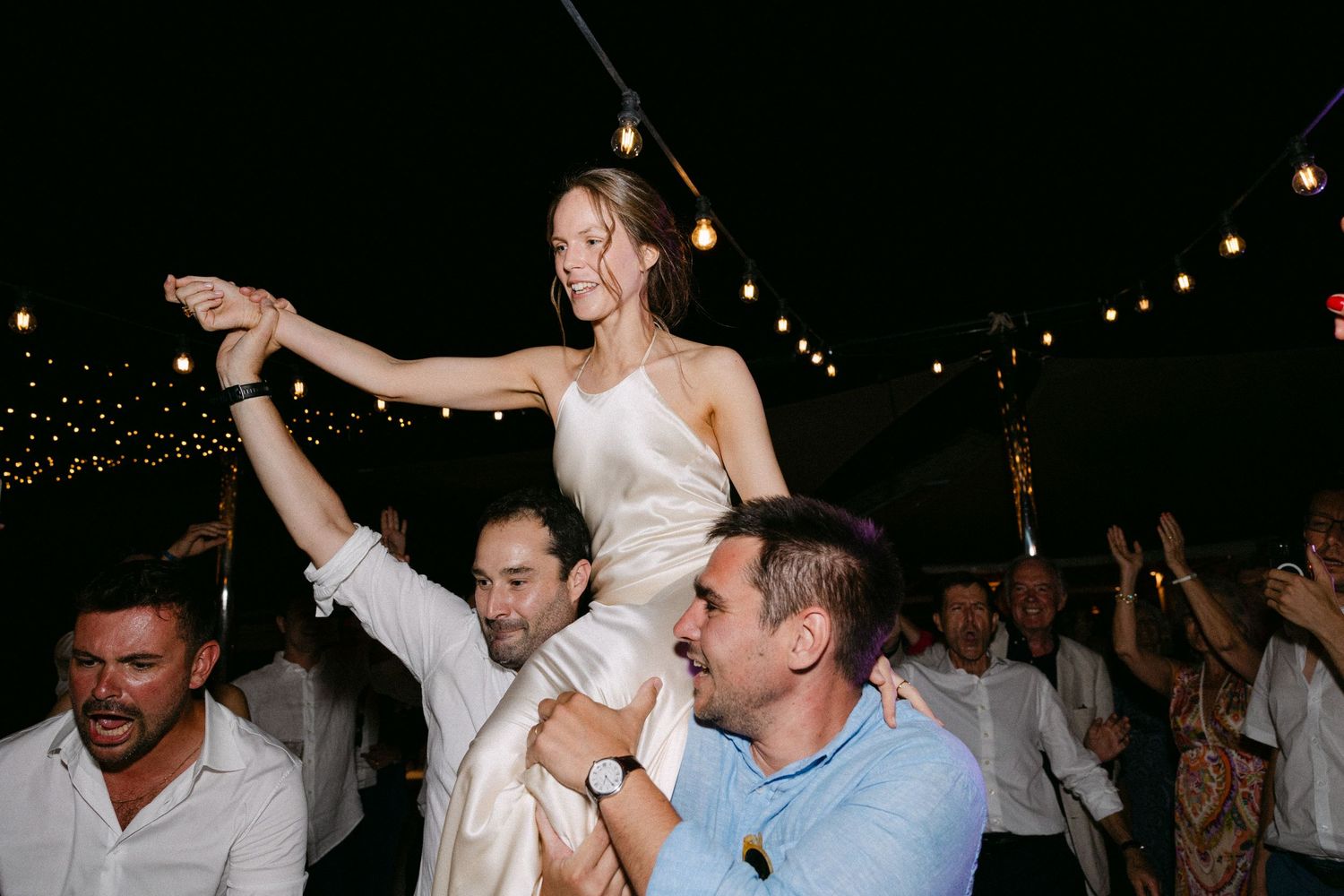 a bride sitting on two wedding guests' shoulder and dancing