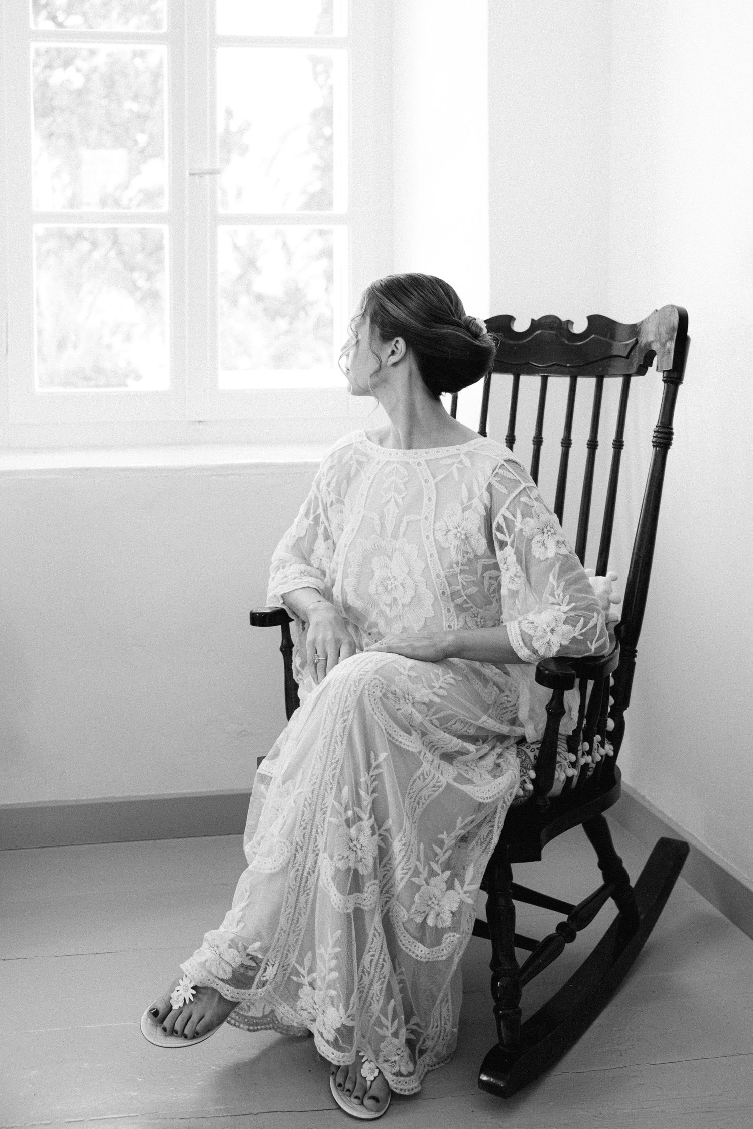 a black and white photo of a bride sitting in a old vintage wooden rocking armchair in a white room and looking out the window