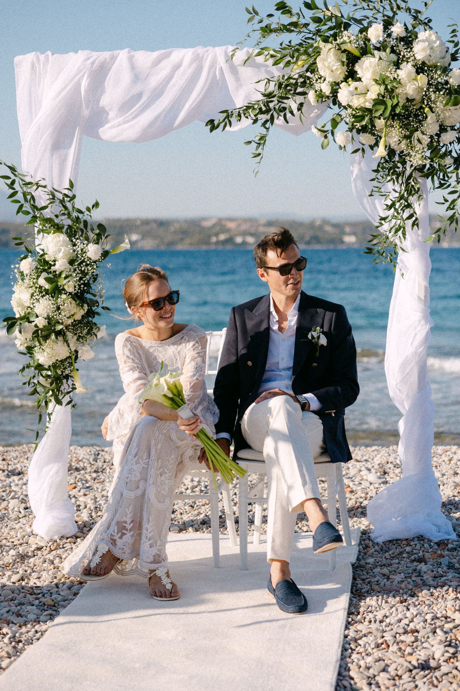 a bride and groom who wear sunglasses sitting in chairs under a drape arch with white flowers during their beach wedding ceremony smiling