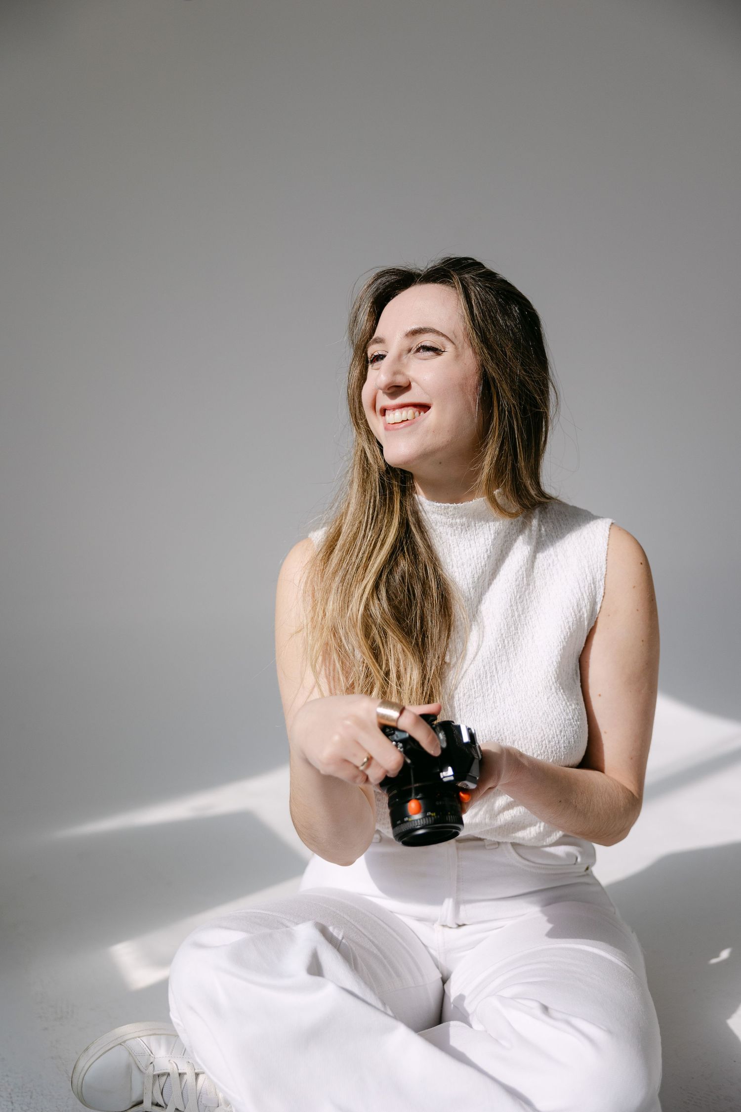 Wedding Photographer Chara Moss' portrait holding a camera, wearing white clothes and smiling against a white background