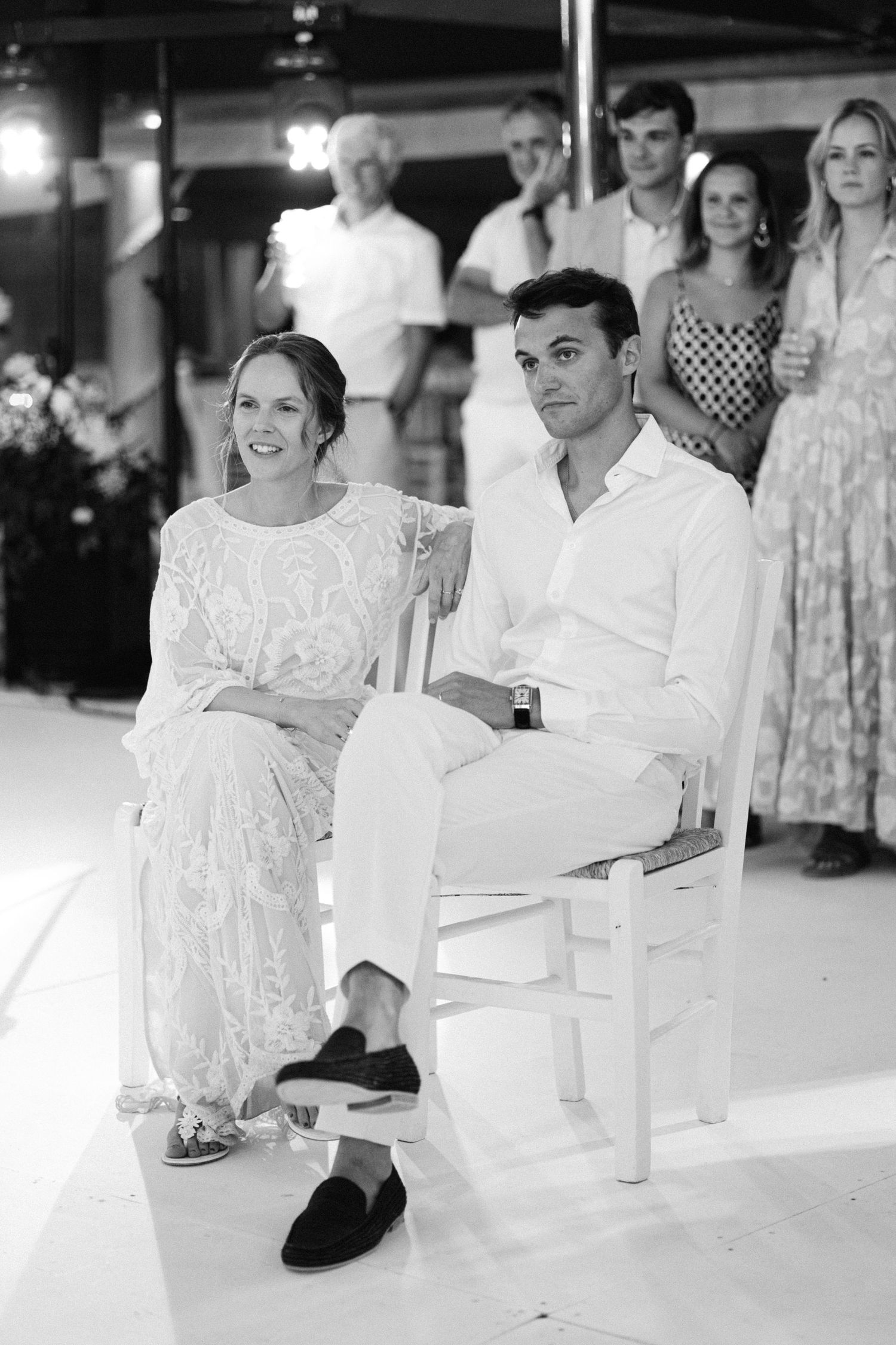 a black and white photo of newlyweds sitting in chairs listening to a speech
