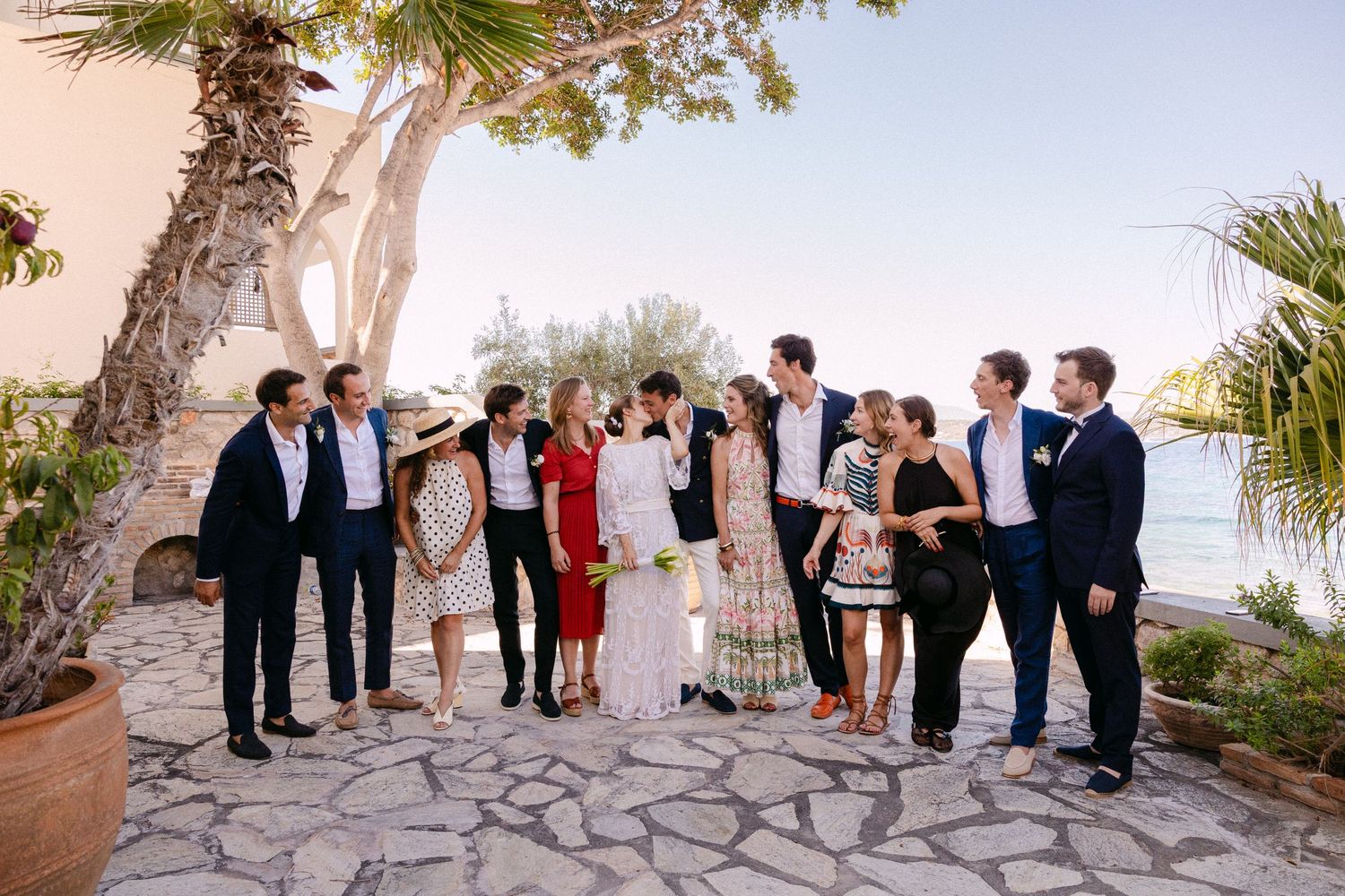a group photo of a bride, groom, groomsmen and other guests looking at each other while the couple is kissing in an open-air summer setting