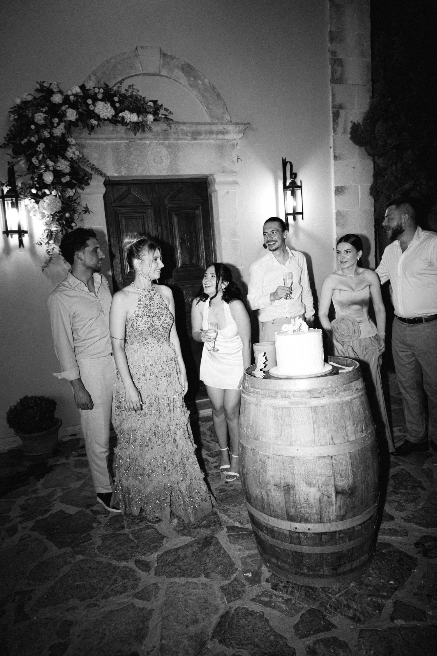 a black and white photo of the couple and wedding guests standing in front of a chapel at night with a cake in front of them and smiling
