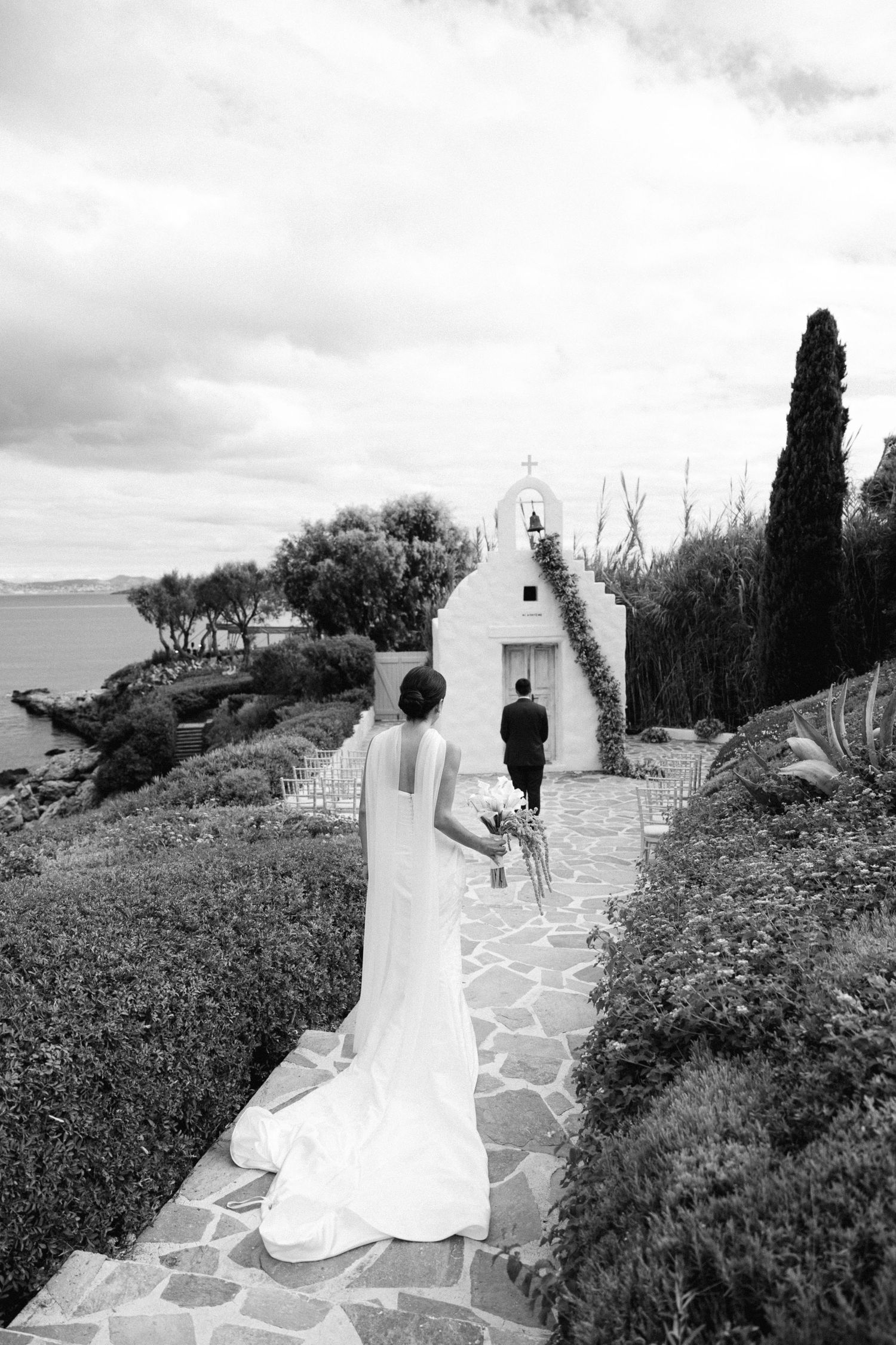 A black and white wedding photo showing the back of a bride walking towards the groom outside a seaside chapel in Athens Riviera Greece