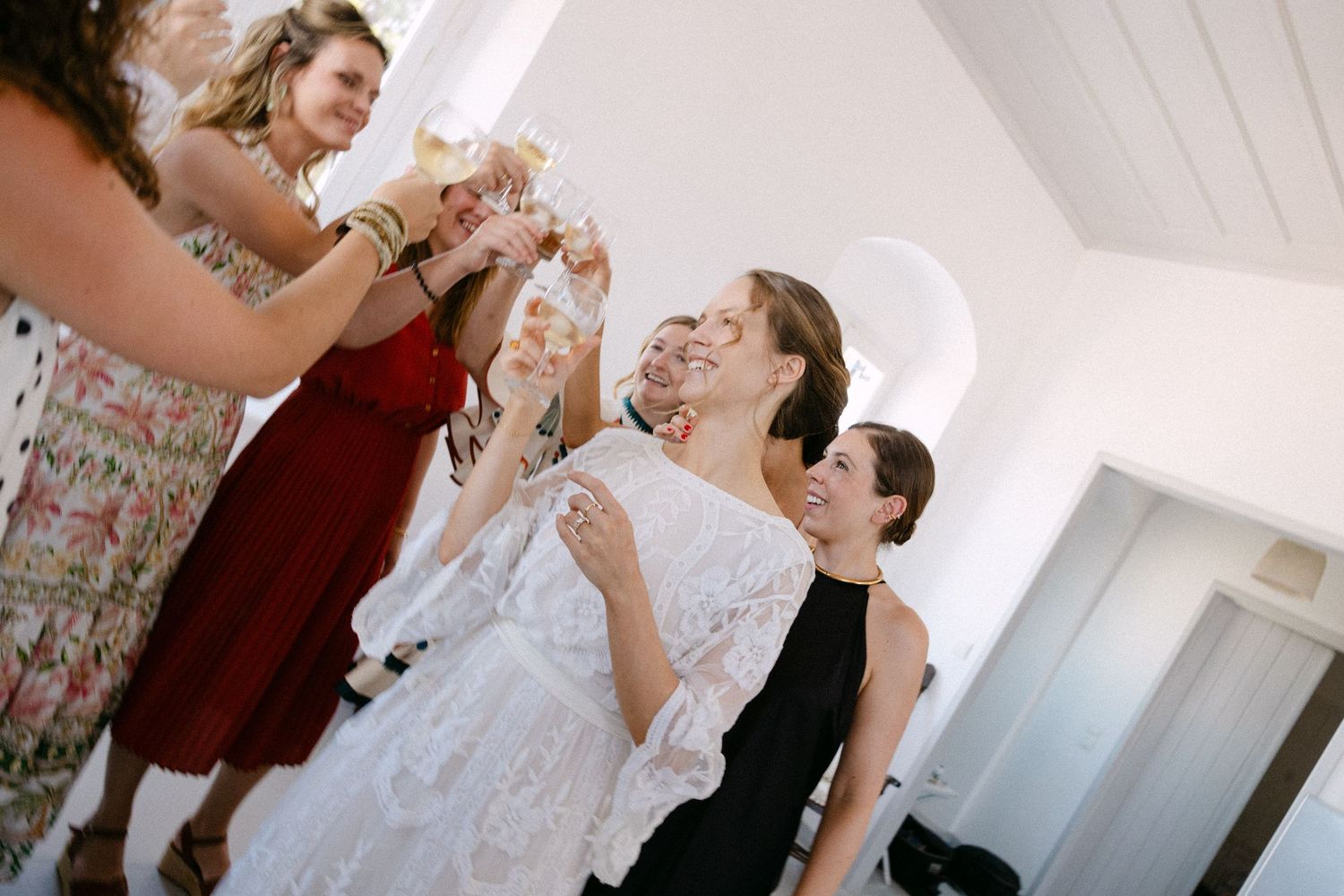 a crooked-horizon photo of a bride clicking glasses with friends in a white summer house during her getting ready