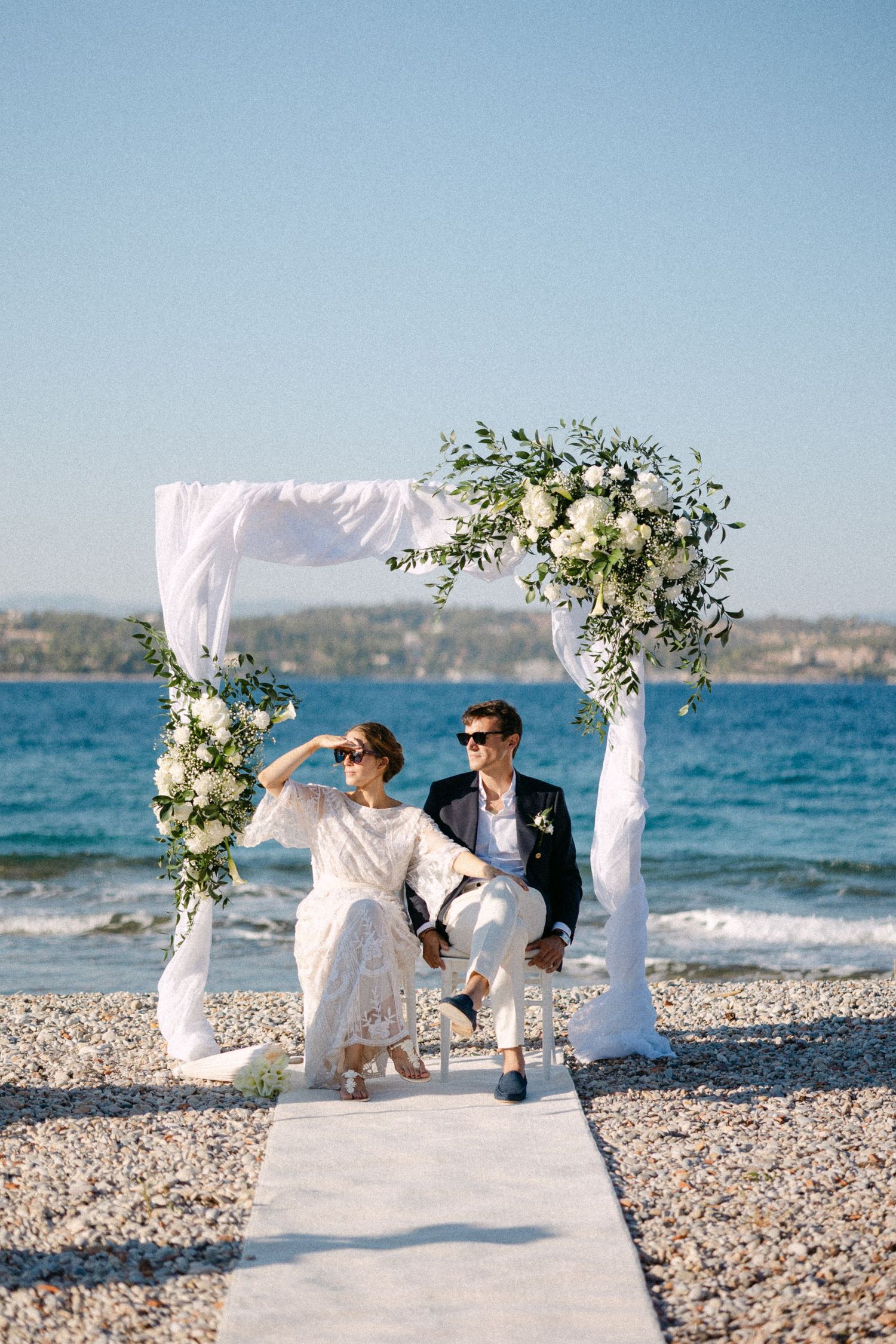 a bride and groom who wear sunglasses sitting in chairs under a drape arch with white flowers during their beach wedding ceremony