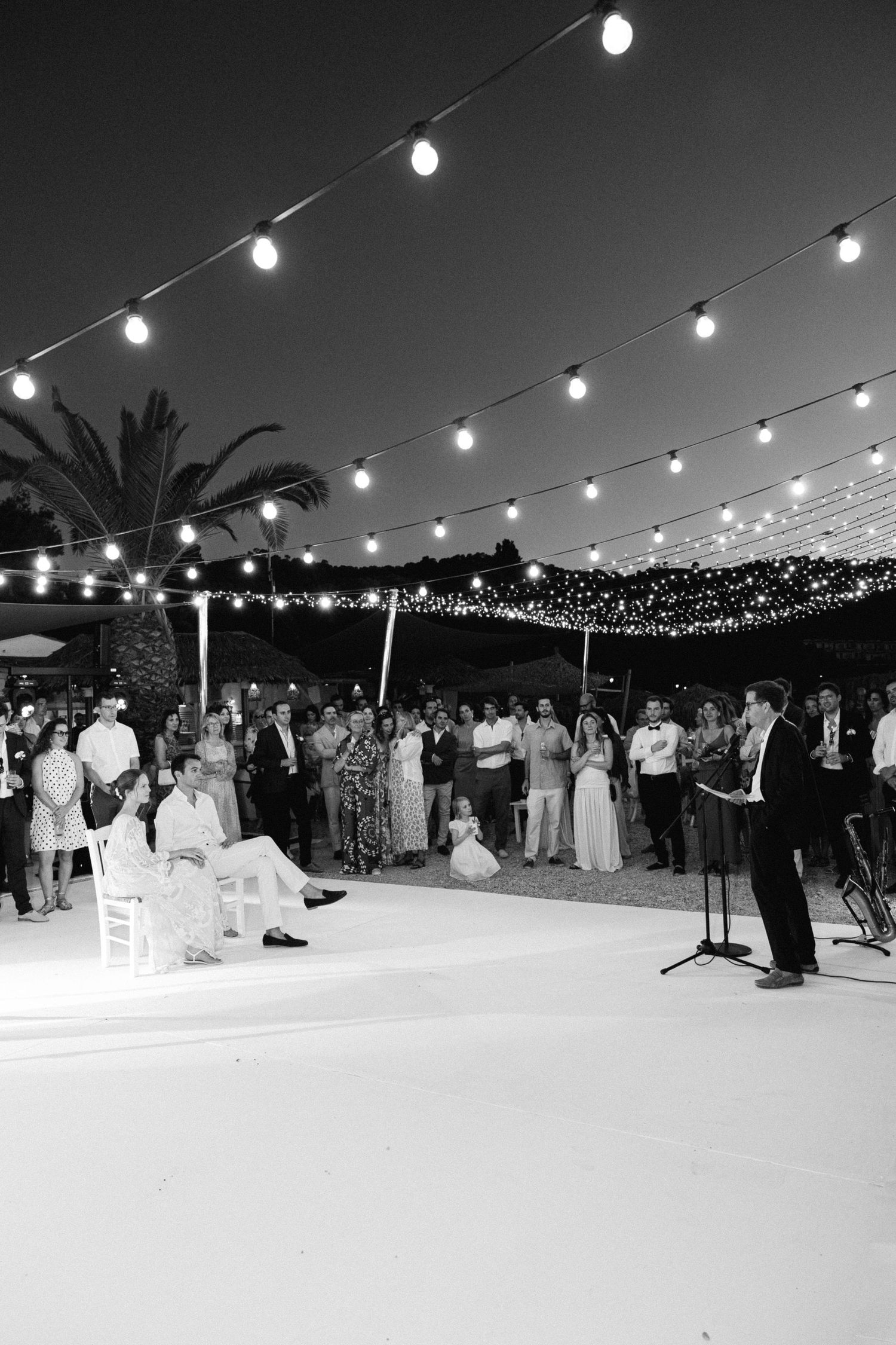 a black and white photo of a wedding couple sitting in chairs opposite the bride's dad who is reading a speech into a microphone with guests