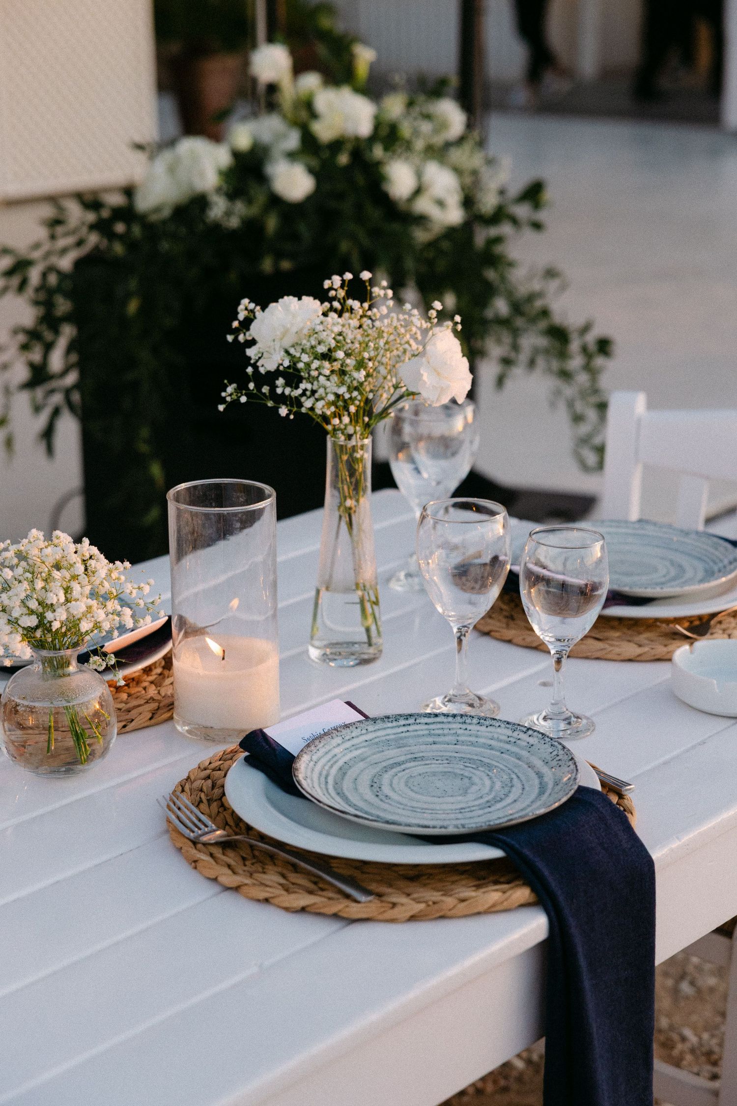 wedding table decorations showing white flowers in a vase, a lit candle and blue plates and glass wines