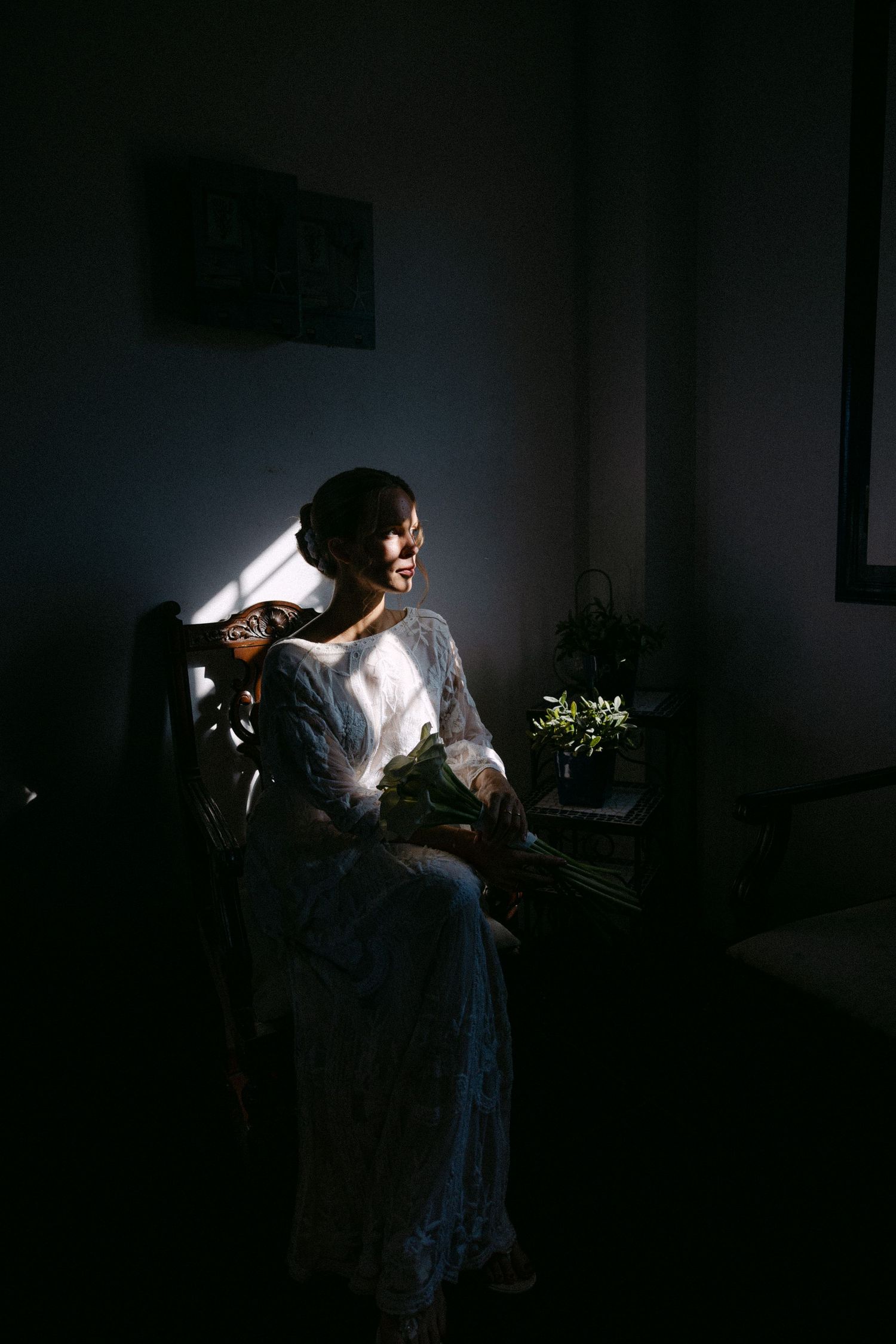 a full-body portrait of a bride being dimly-lit from window light while sitting in an armchair, and looking outside the window
