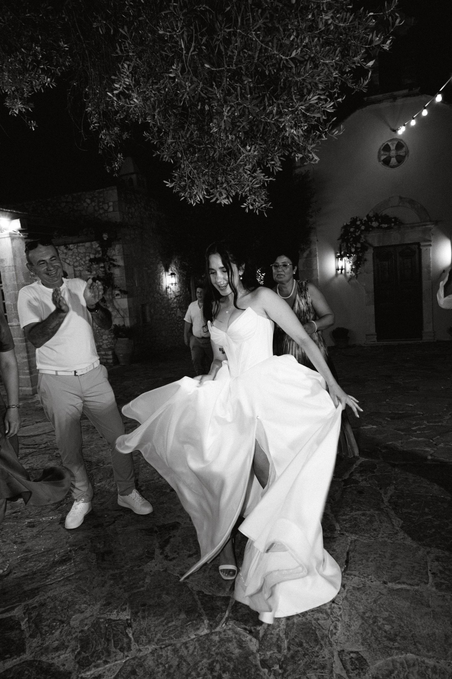 a black and white photo of a bride dancing and playing with her wedding dress while people are cheering in the background