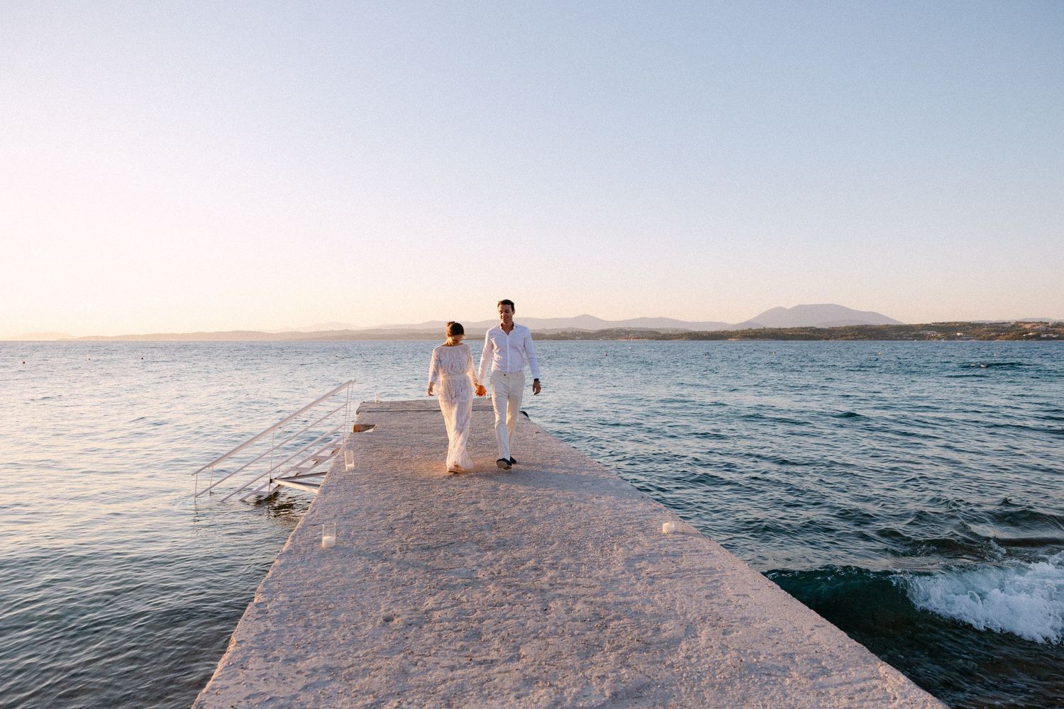 a bride and groom walking and holding hands at a dock at sunset