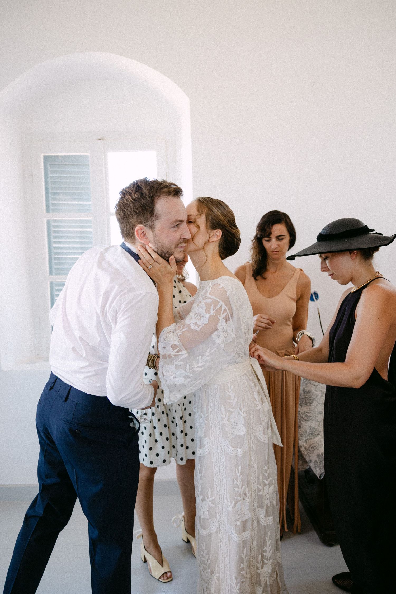 a bride kissing her brother on the cheek while a friend wearing a black hat is fixing her dress from the back