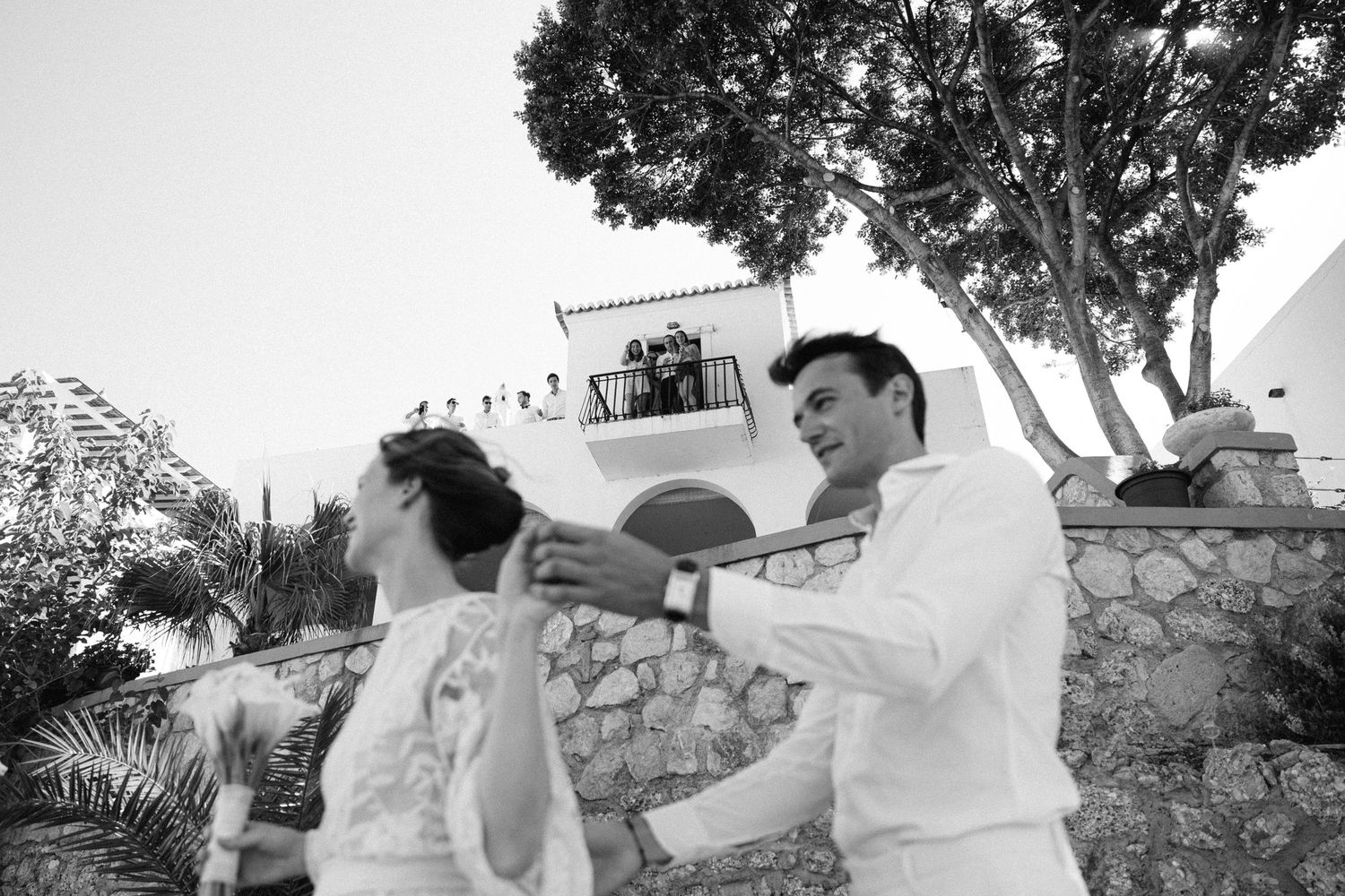 a black and white photo of a bride and groom dancing white a house visible in the background where guests are cheering