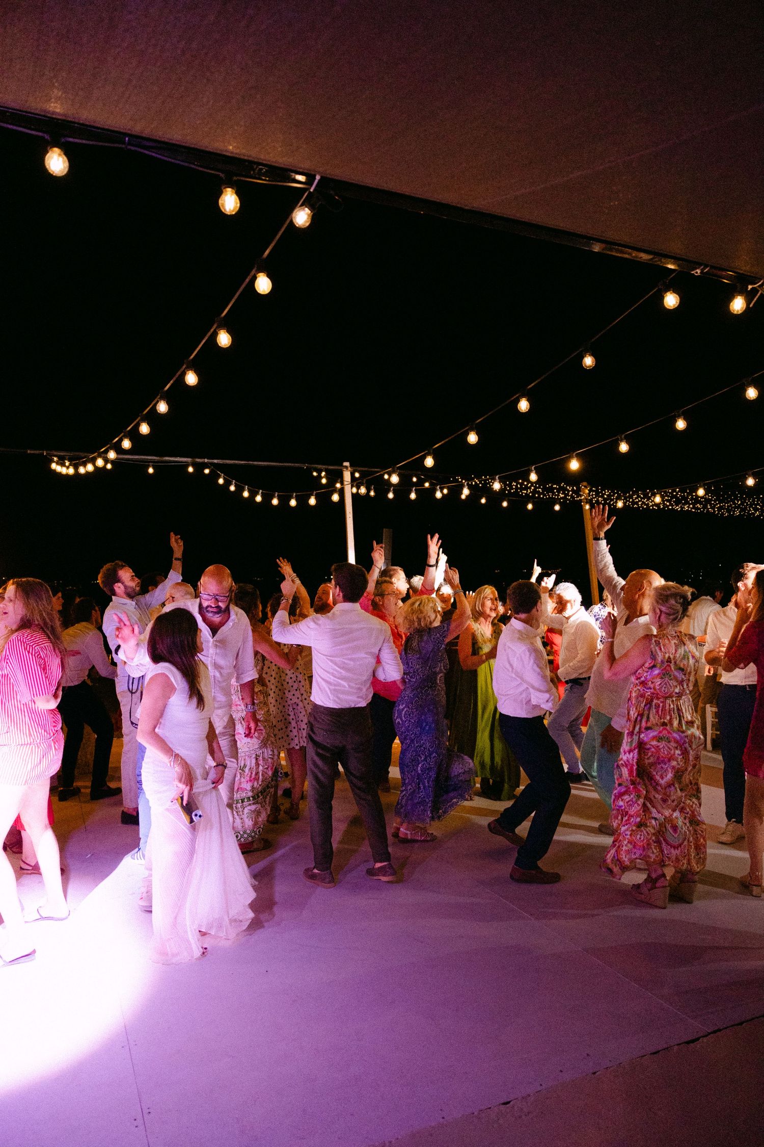 a wide party photo of guests dancing with hands up in the air in an open-air wedding dance party