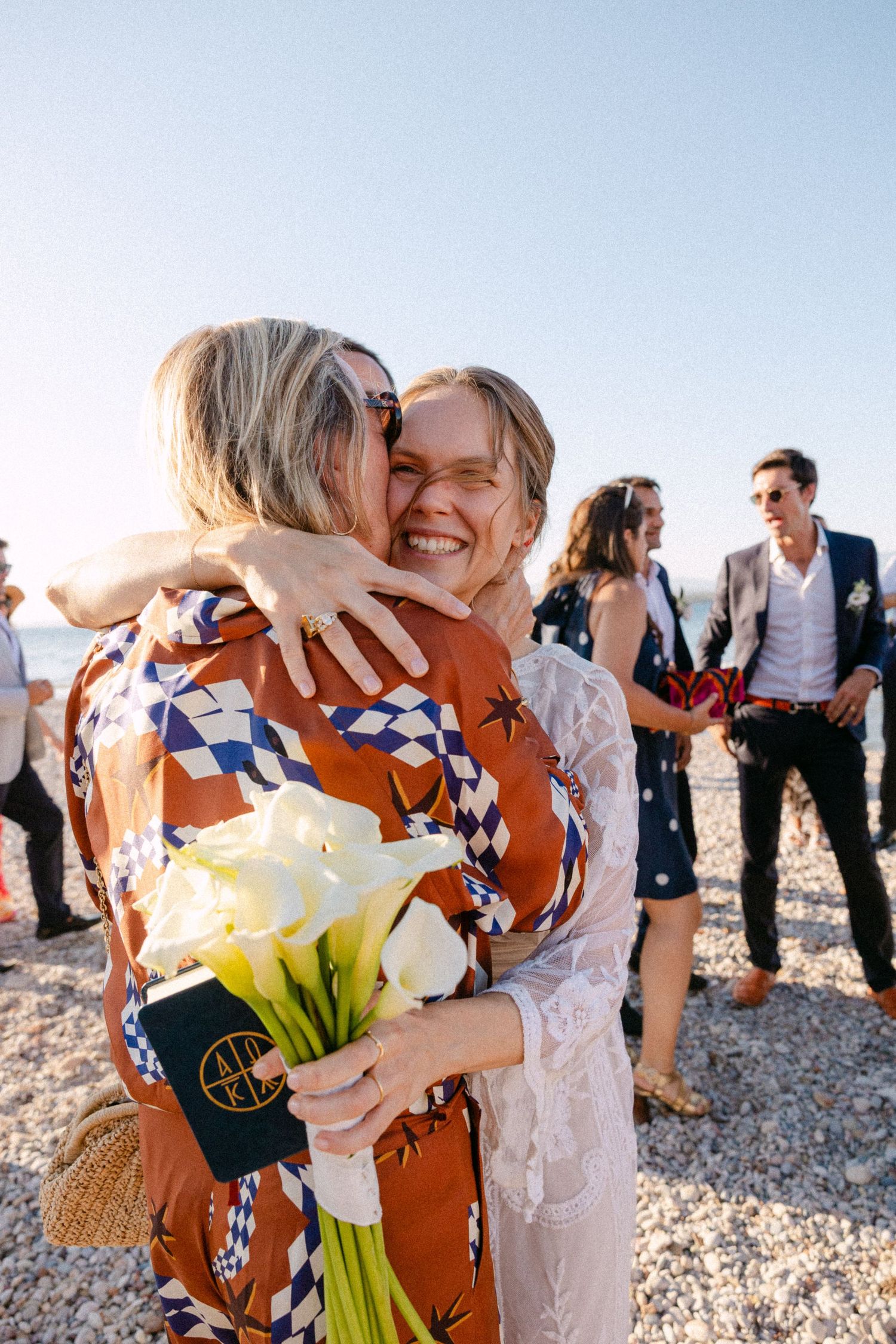 a bride hugging a guest among other guests at a beach at afternoon, holding a white Lillies bouquet
