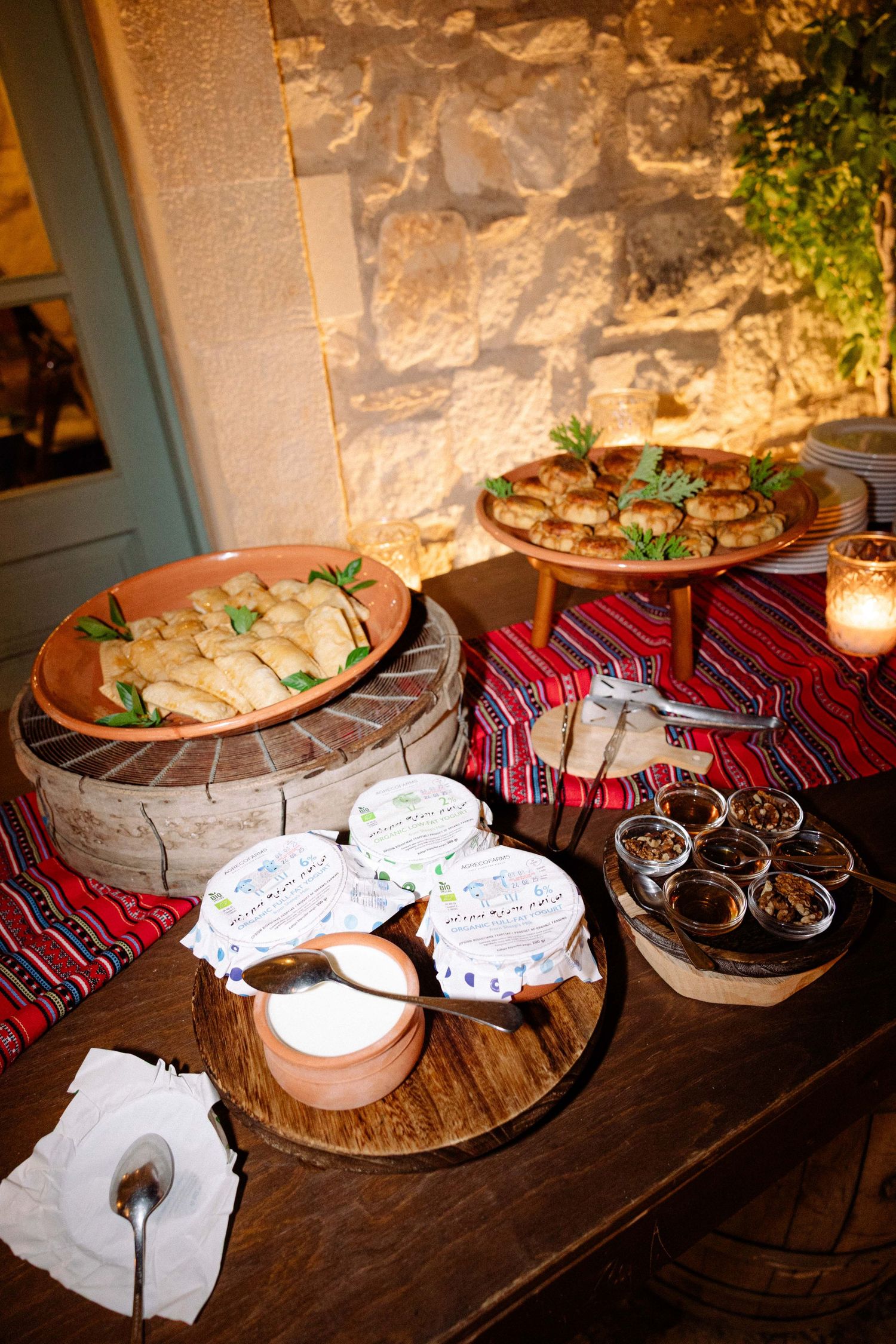 a buffet of greek cuisine snacks like greek yogurt, pies, marmalade, set in a wooden table against a stone wall backdrop