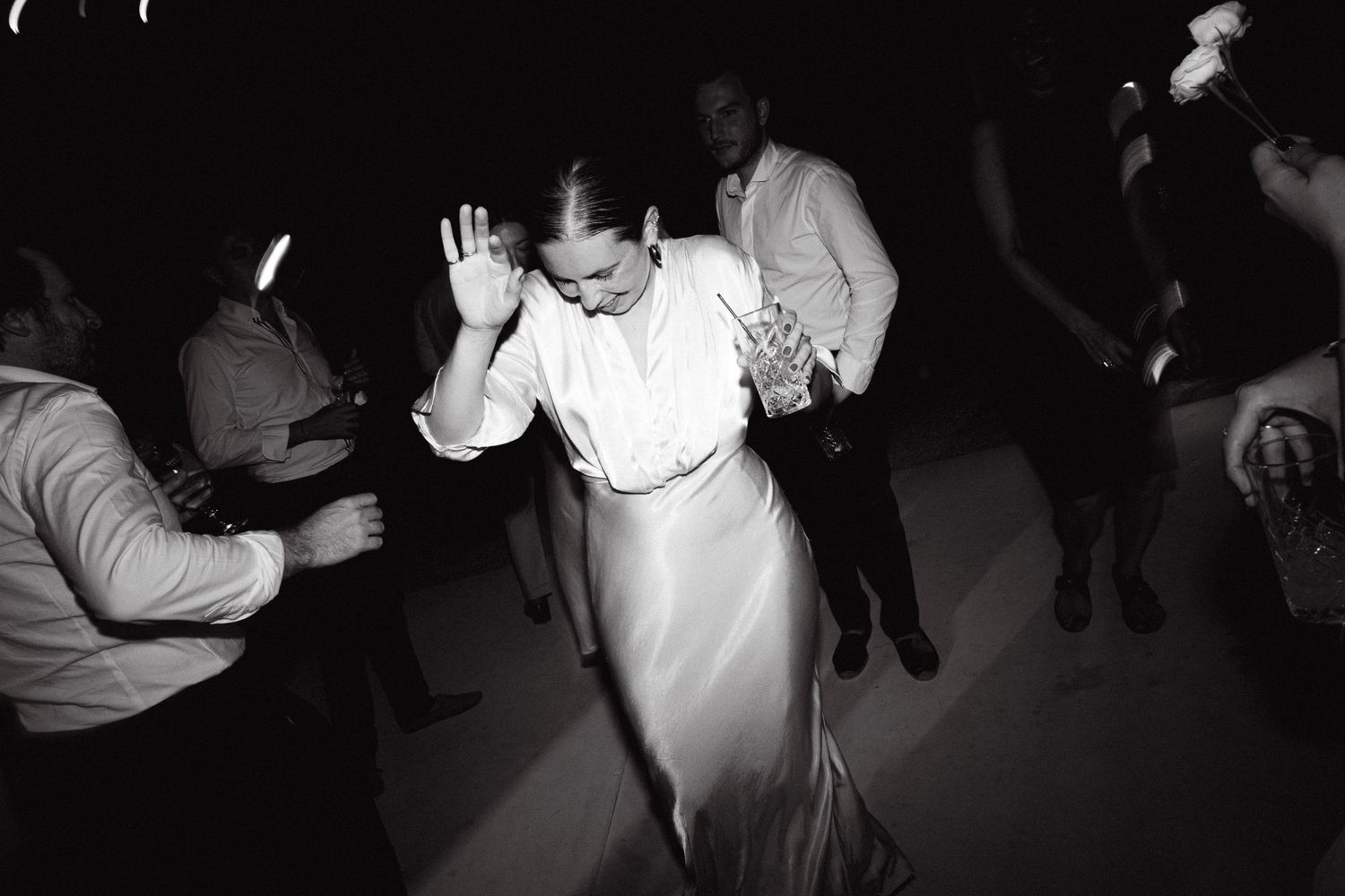 a black and white photo of a wedding guest dancing with hands up in the air holding a drink with a straw