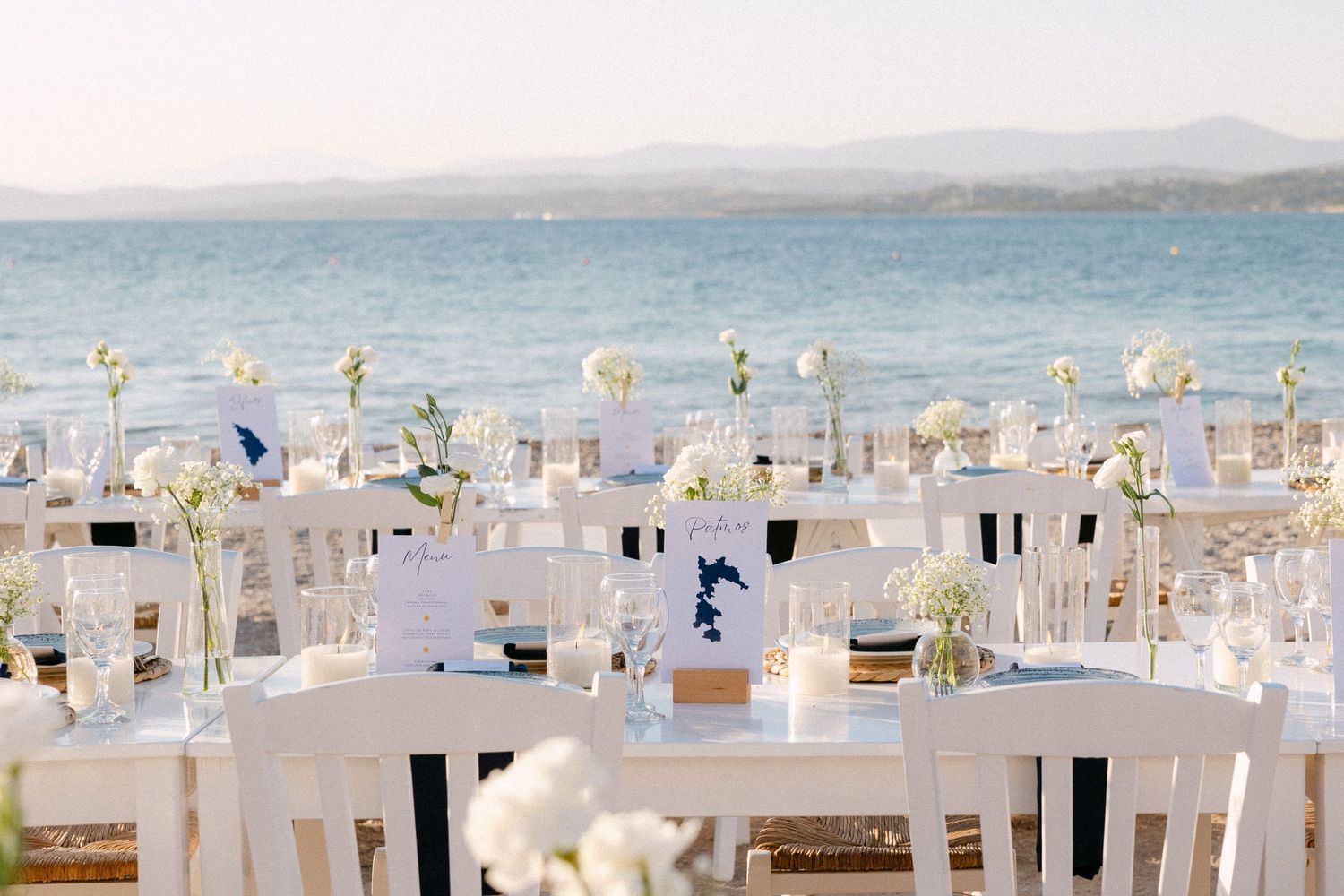 wedding table decorations showing white flowers in a vase, a lit candle, blue plates, glass wines and white chairs with a sea background