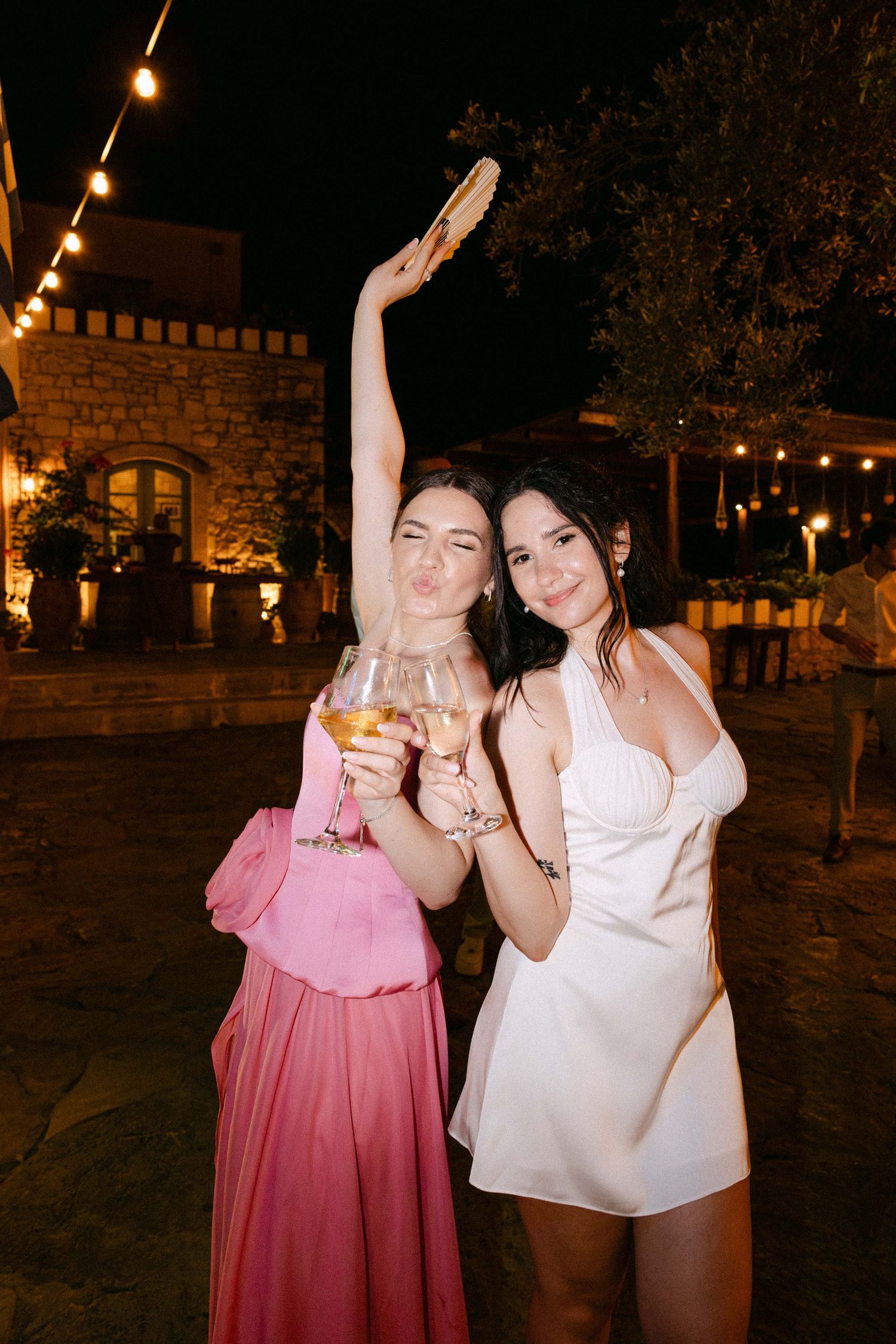 a bride and her friend who wears a pink dress are dancing outdoors at night while holding drinks and posing for the camera