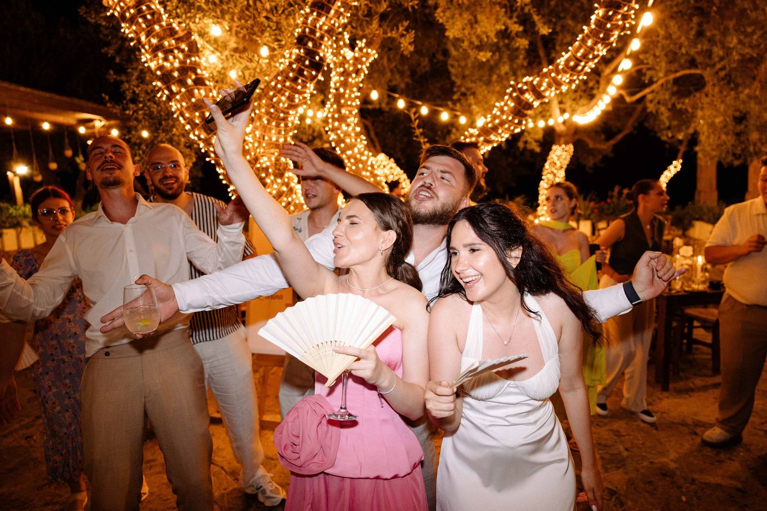 a bride and wedding guests dancing and holding fans at night surronded by trees with fairy-lights