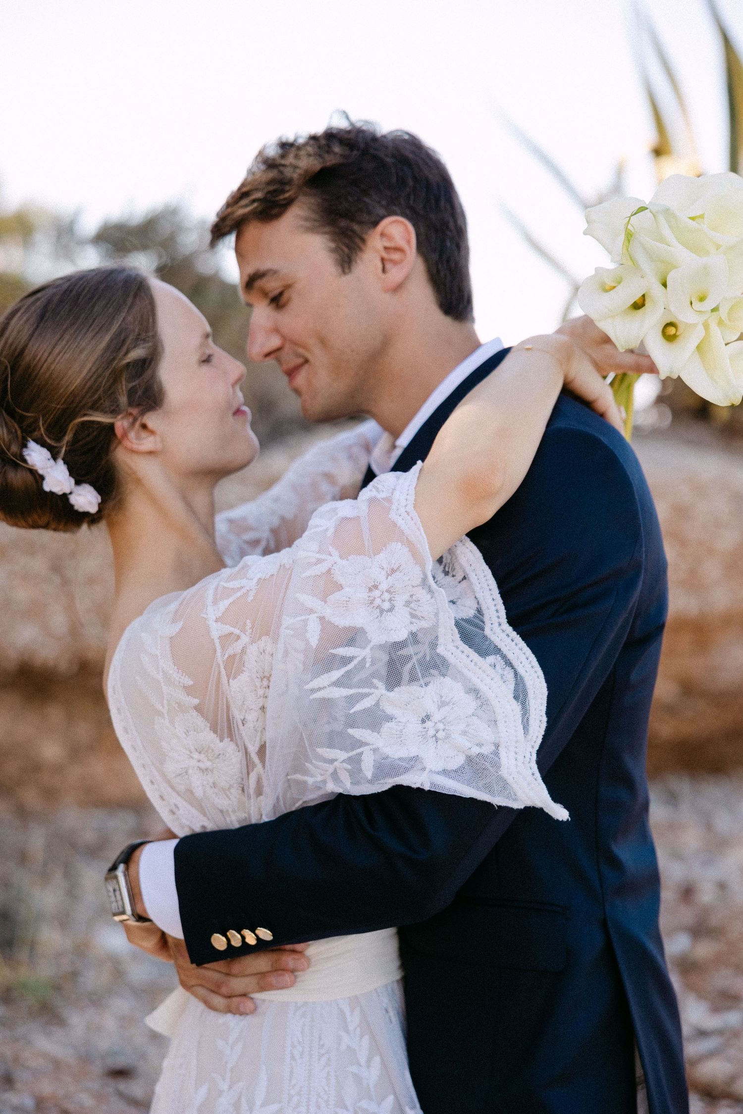 a bride has her hands wrapped around a groom and they look each other in the eyes lovingly and smiling - the bride is holding a bouquet