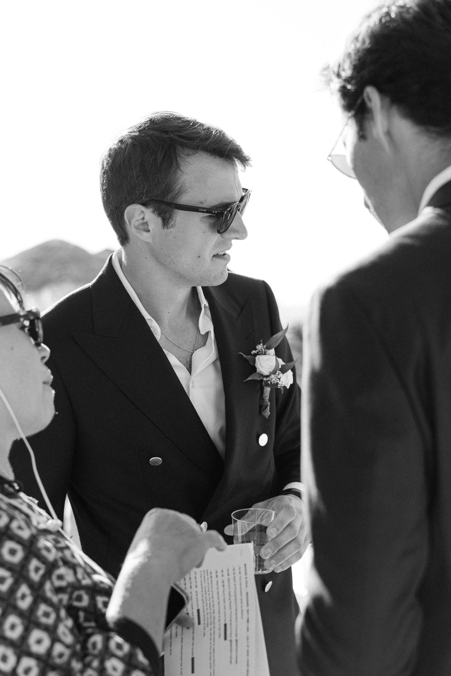 a black and white groom portrait of him talking to people and wearing shades and a black suit and white shirt