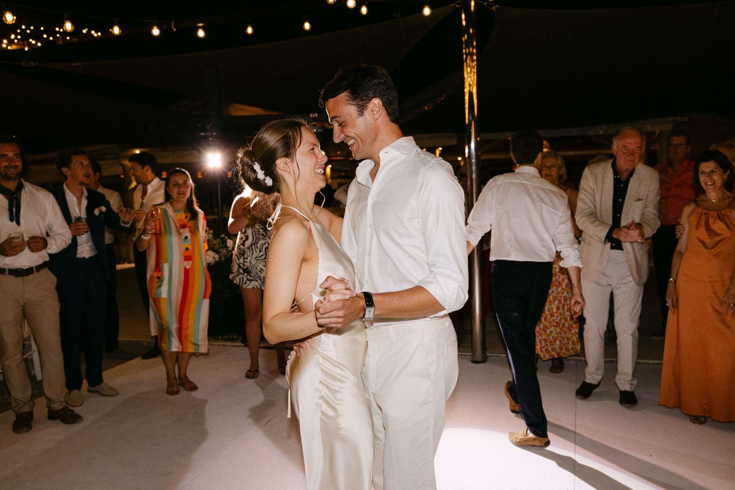 a bride and groom slow dancing with each other and smiling with the guests as backdrop