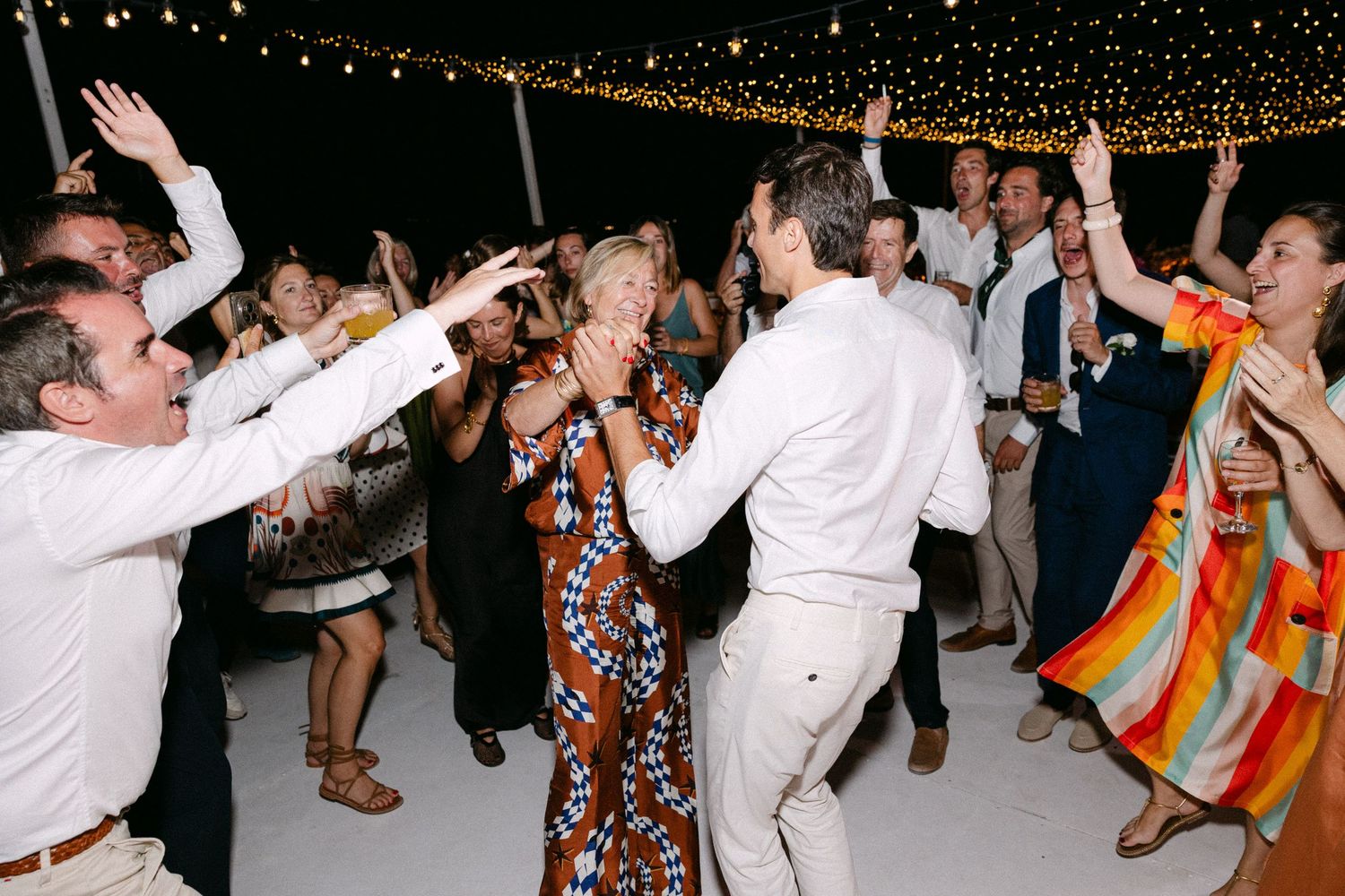 a groom and his mum dancing together under carnival lights as surrounding guests are cheering