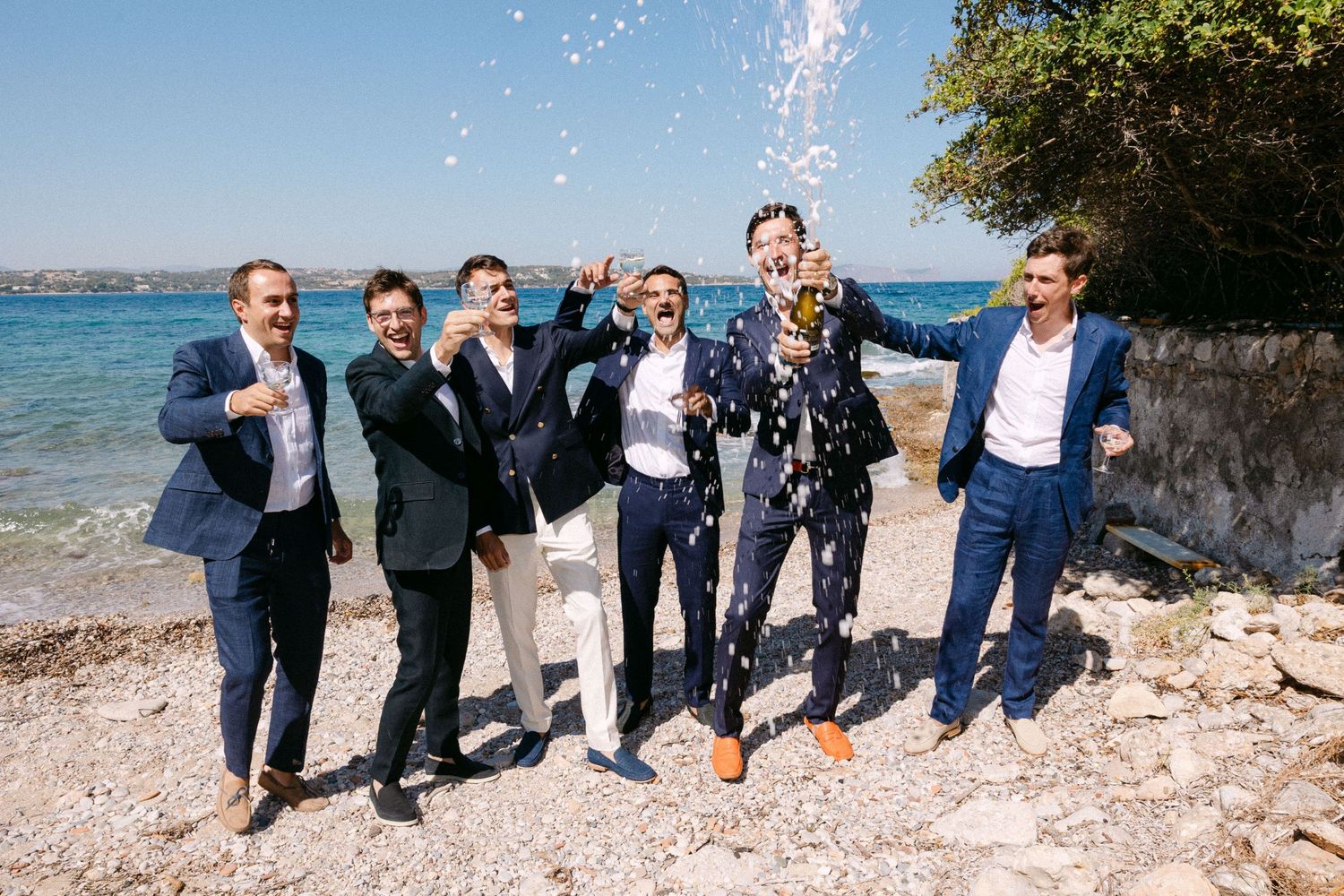 a groom and five groomsmen dressed in navy blue suits in a beach - one of them is opening a splashing champagne and they are all cheering