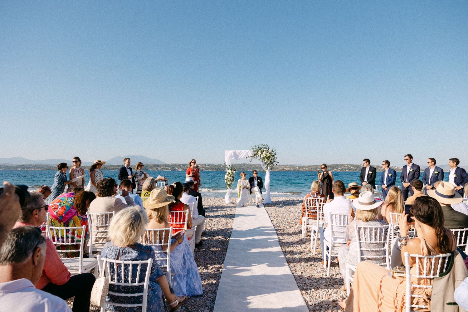 a wide photo of a beach summer wedding ceremony taken from the middle of the aisle, where the bride and groom are sitting