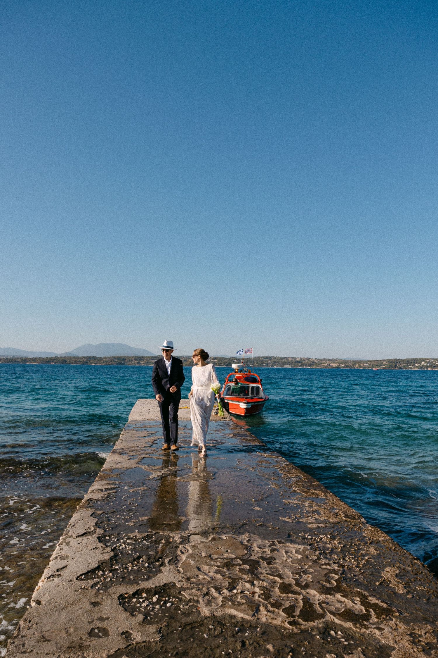 bride and her dad walking in a dock, while the bride is waving and a red boat is visible in the background
