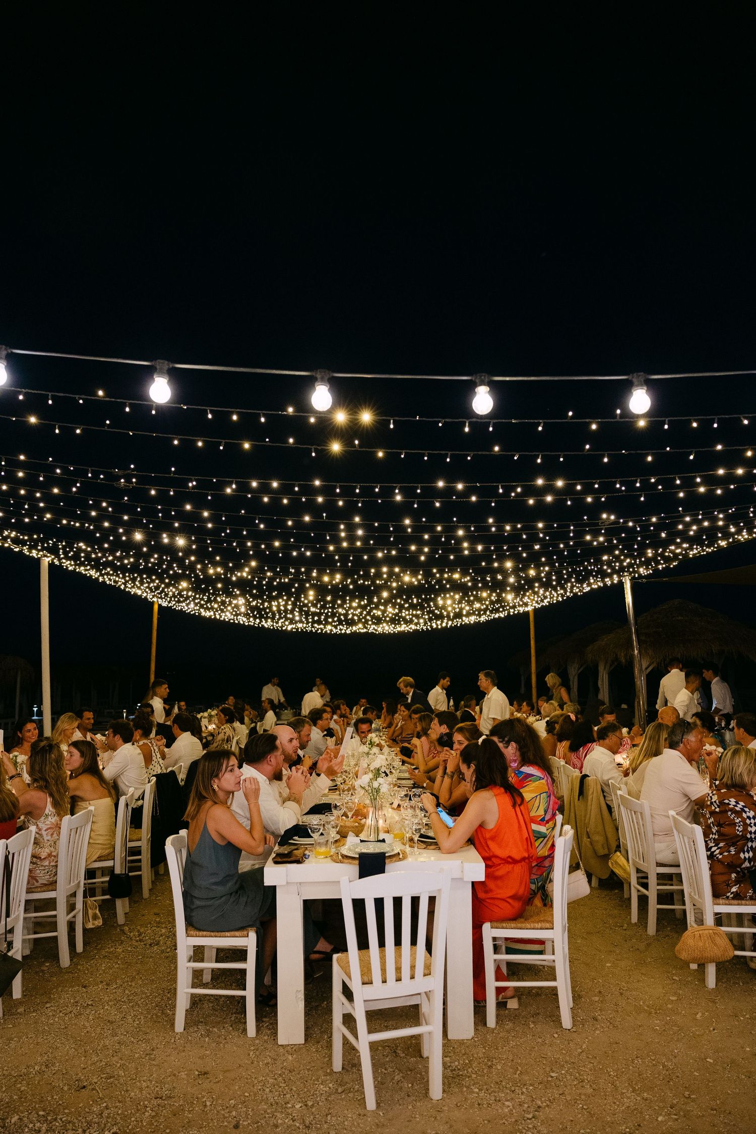 a wide photo of an open-air wedding dinner in a beach bar in spetses - there are three wide tables under carnival lights