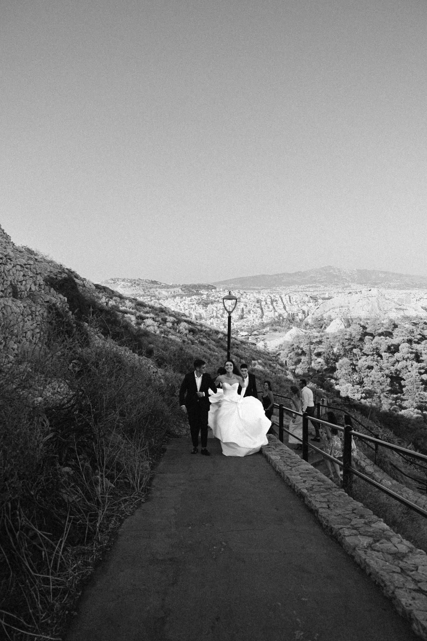 a black and white wide photo of a bride being held by her brother and surrounded by relatives walking in a city view background