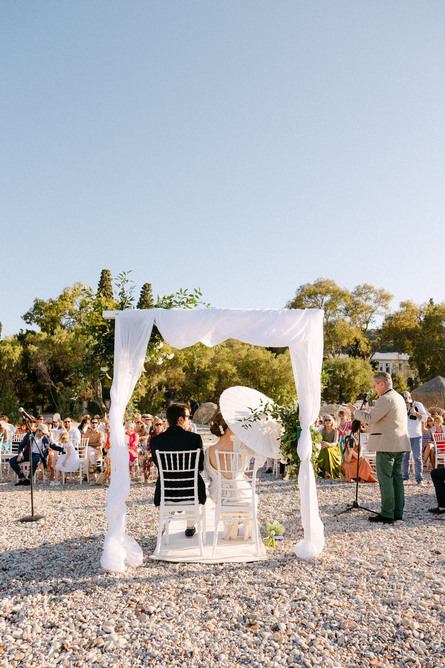 a bride and groom sitting in chairs under a drape aisle during their wedding ceremony. The bride is holding a sun umbrella.
