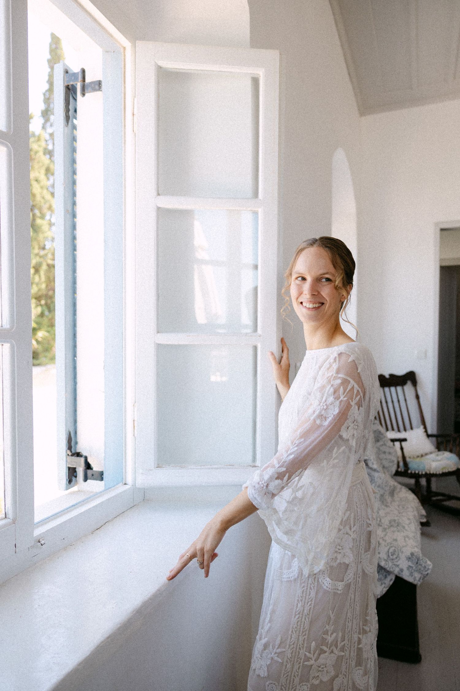 a bride with an up hair do and a lace long-sleeve wedding dress opening a window inside a White island summer house, smiling at the camera