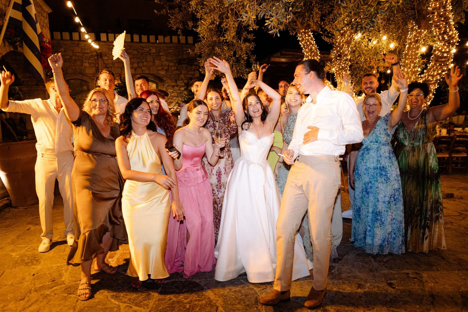 about 30 wedding guests, a bride and a groom are taking a group photo cheering and laughing candidly at a wedding night reception outdoors