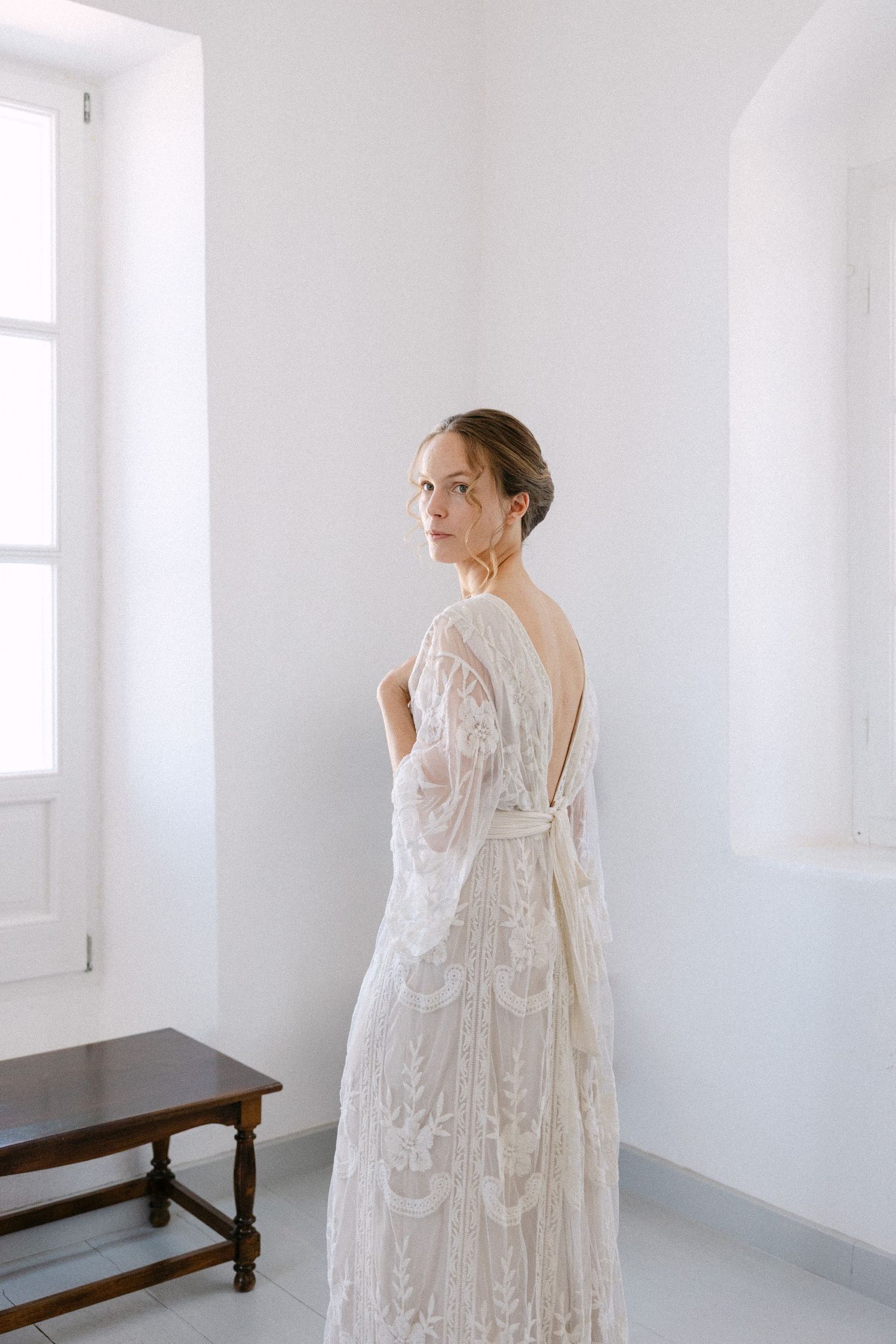 a bride with an up hair do and a lace long-sleeve wedding dress standing in a white indoors background, with the back half-turned