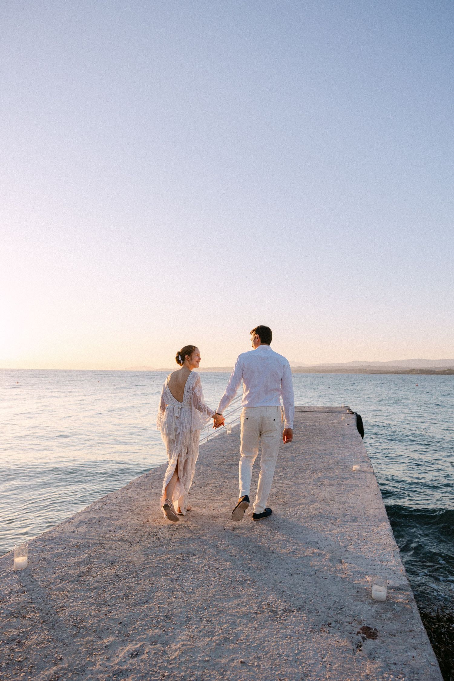a bride and groom walking and holding hands at a dock at sunset