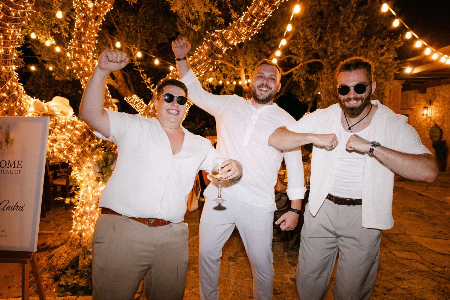 three people dressed in white are dancing outdoors at night surrounded by trees with fairy-lights of warm lighting