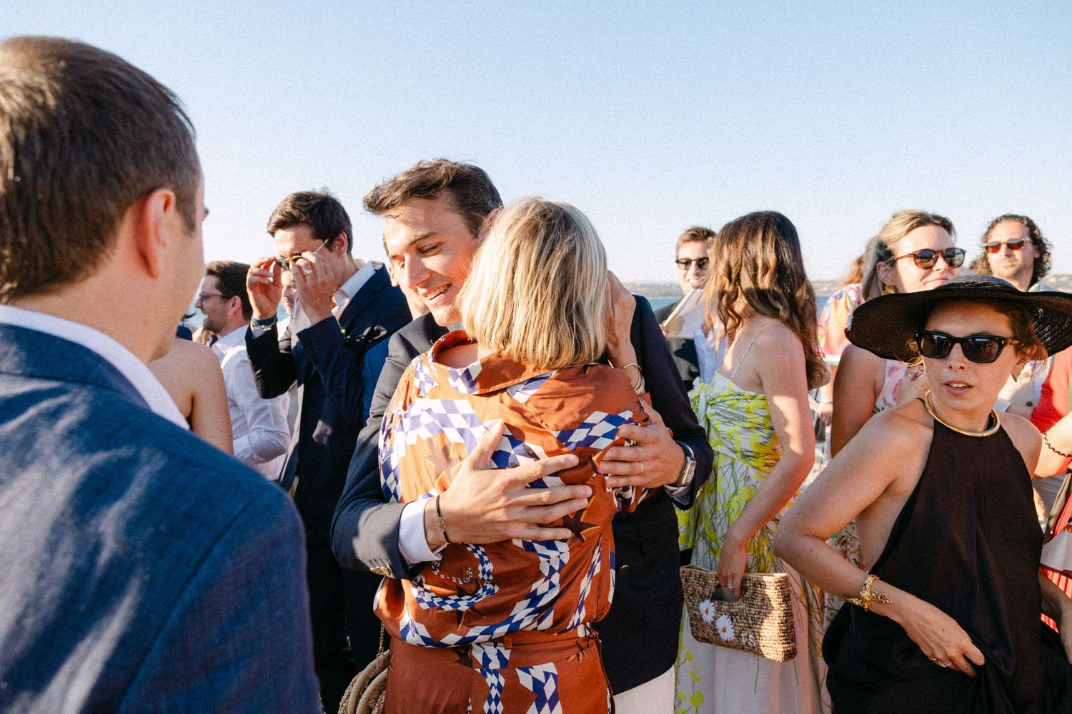 a groom hugging a guest among other guests at a beach at afternoon